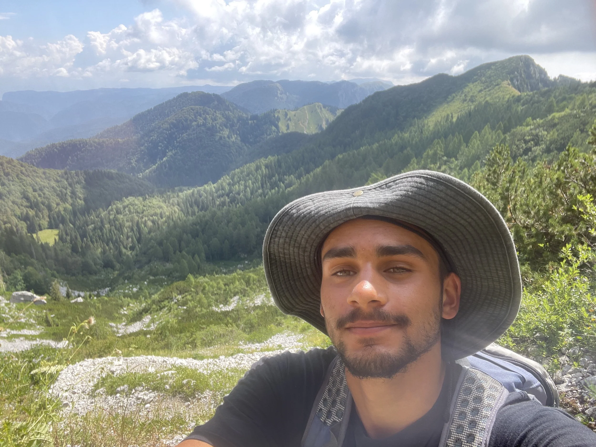 A person wearing a wide-brimmed hat and a backpack takes a selfie during a hike in a lush mountainous area with green forests and a cloudy sky.