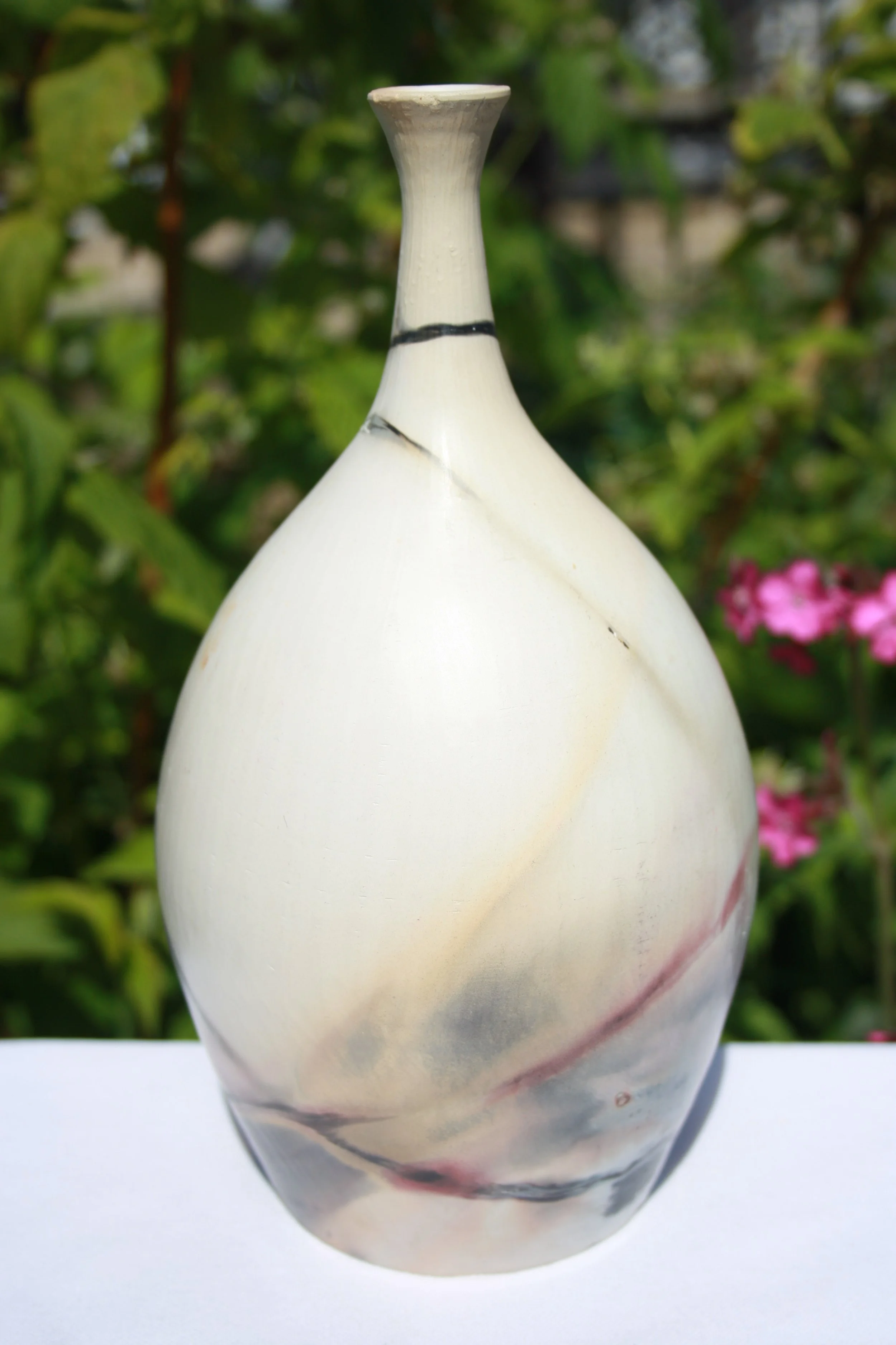Close-up of a decorative ceramic vase with a narrow opening, featuring swirling black, red, and gray patterns on a cream-colored background, outdoors with green foliage and pink flowers in the background.