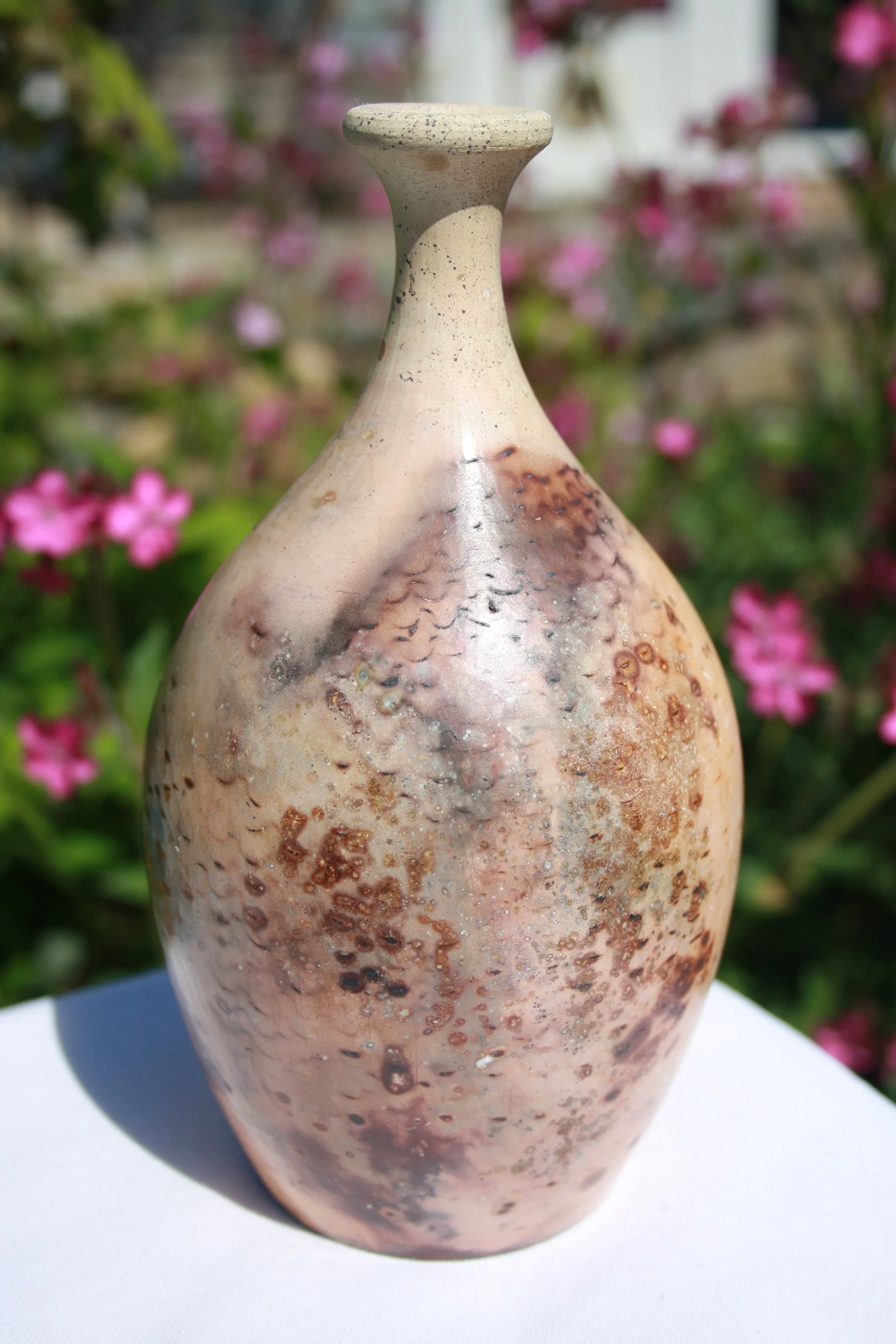 Close-up of a tall, ceramic vase with a speckled, textured surface, positioned on a white surface against a colorful, blurred floral background.