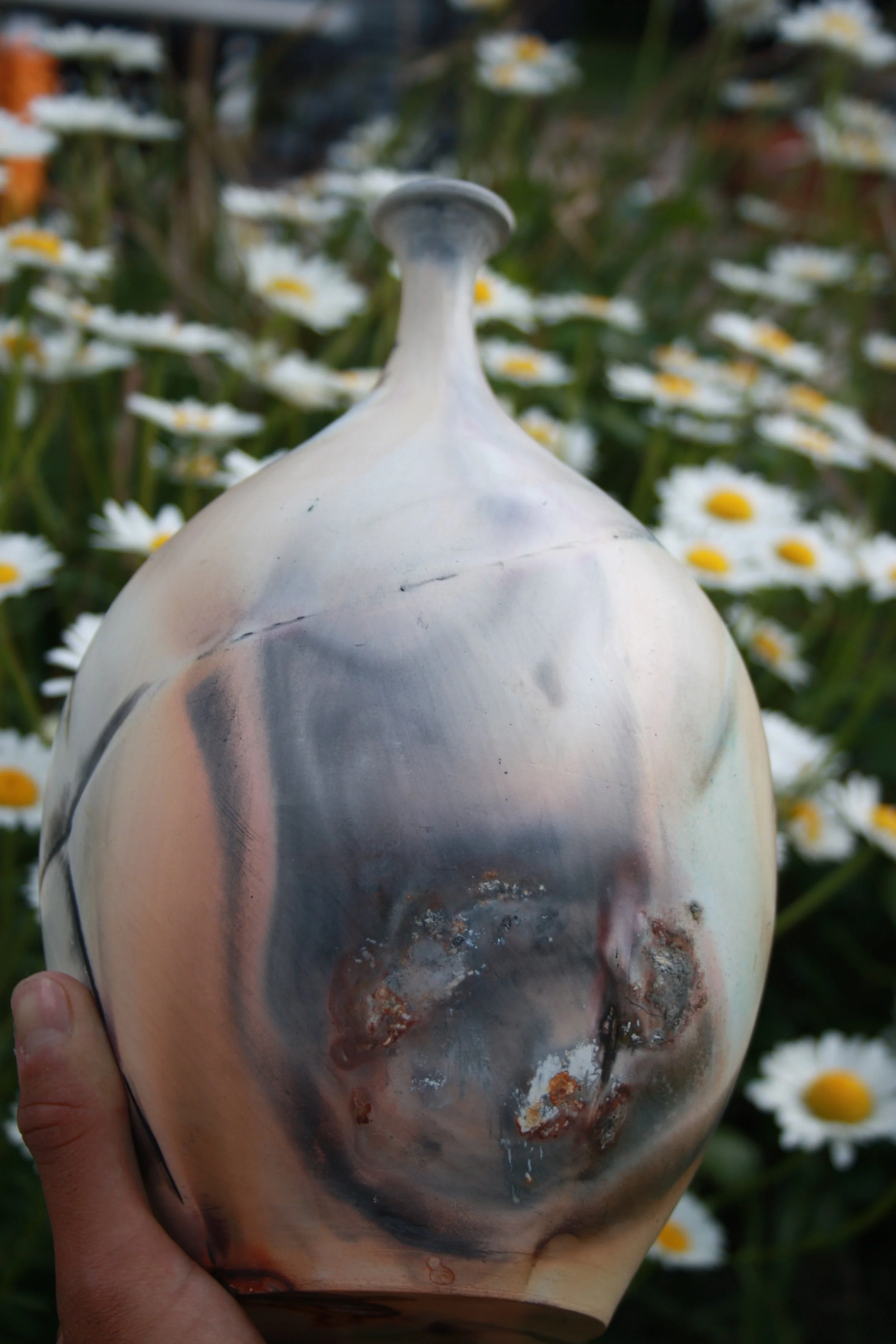 Close-up of a  ceramic vase with dark streaks and rust-like spots, held in a person's hand, with white daisies in the background.
