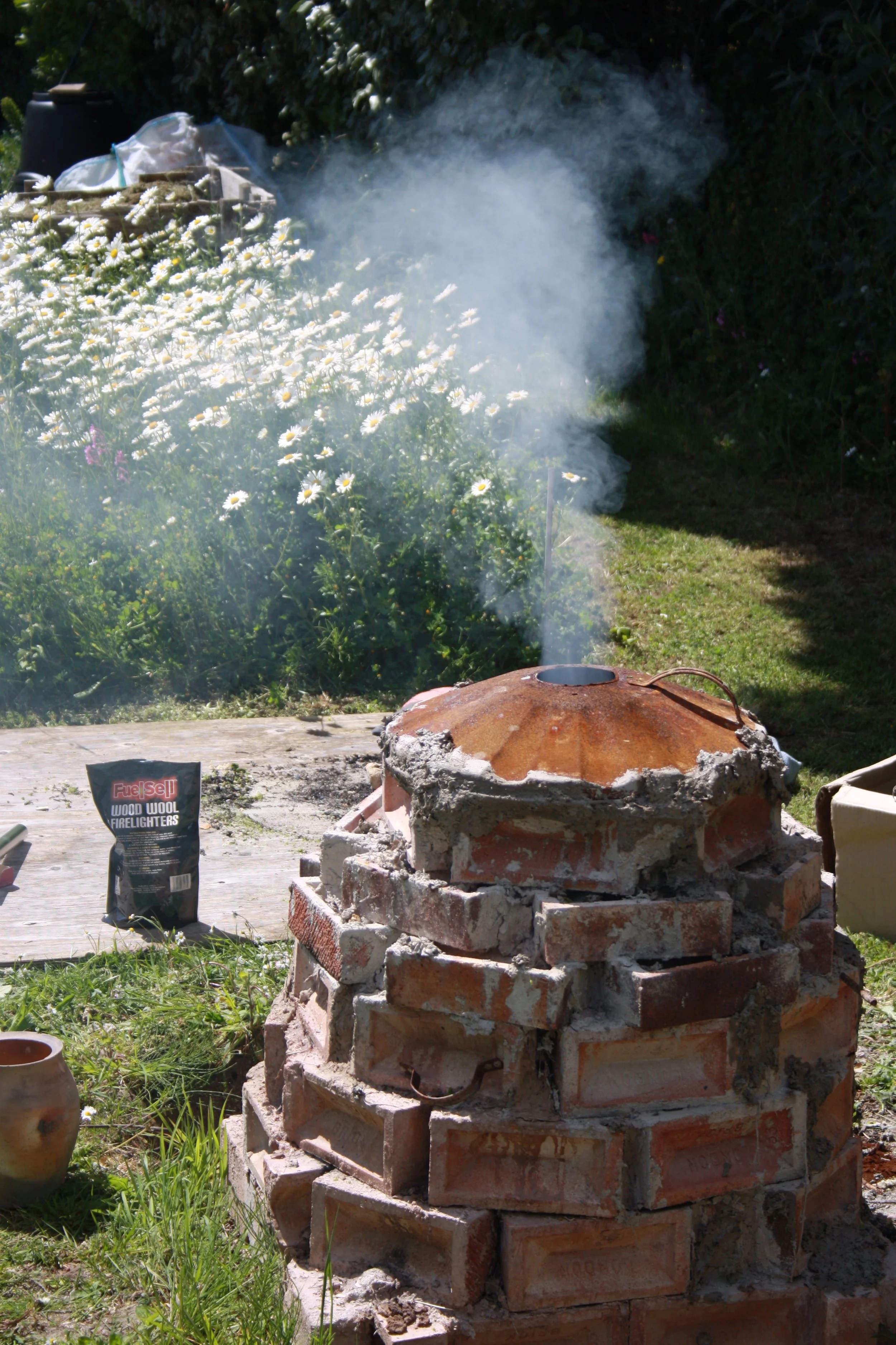 A brick outdoor fireplace with ashes and smoke emerging from the top. There are daisies and gardening supplies nearby, including a bag labeled 'Wood Wool Firelighters'.