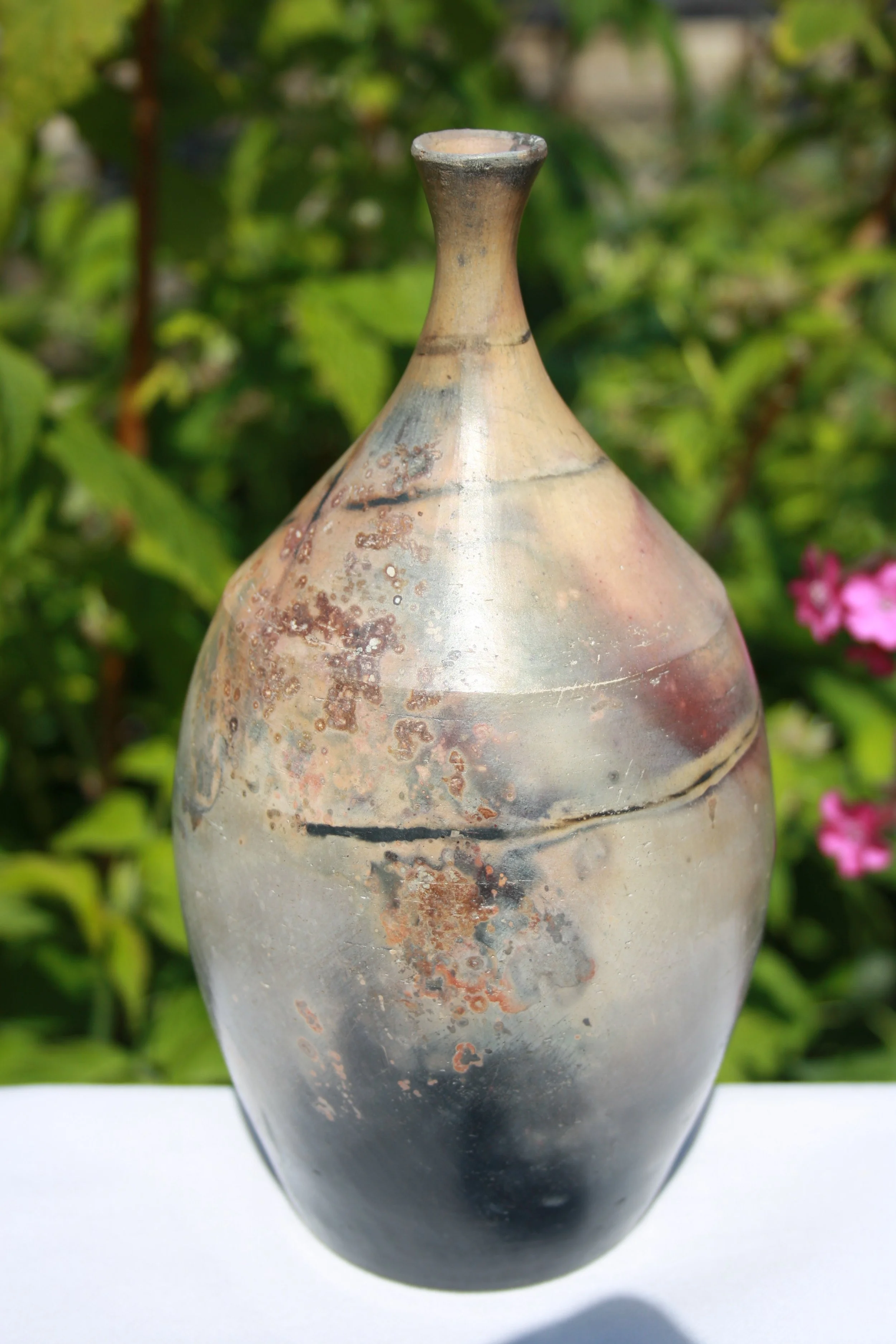 Close-up of a decorative vase with a tapered neck and mottled beige, brown, and gray pattern, set against a green leafy background.