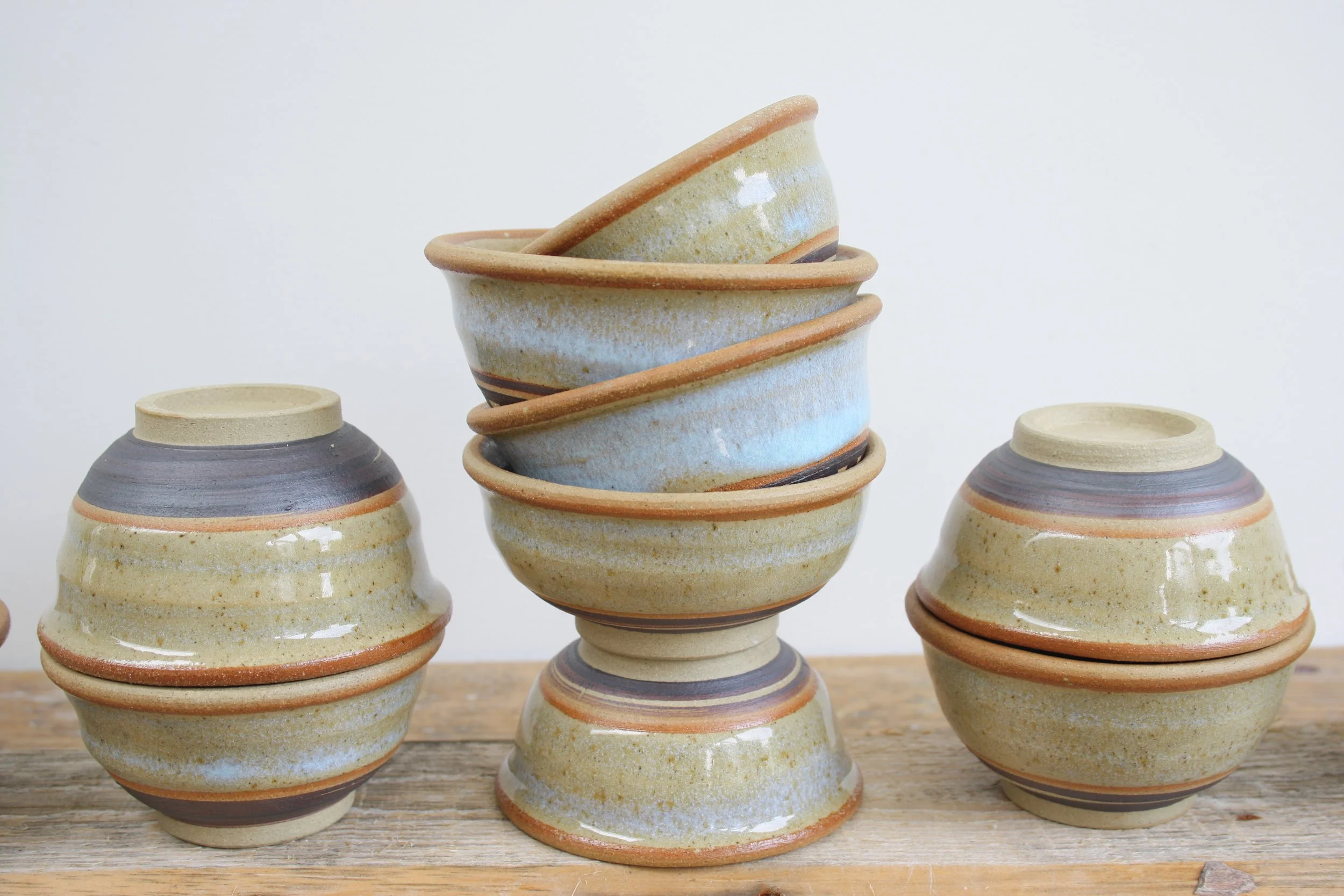 Stack of six handmade ceramic bowls with earthy tones and horizontal stripe patterns, placed on a wooden surface against a plain white background.