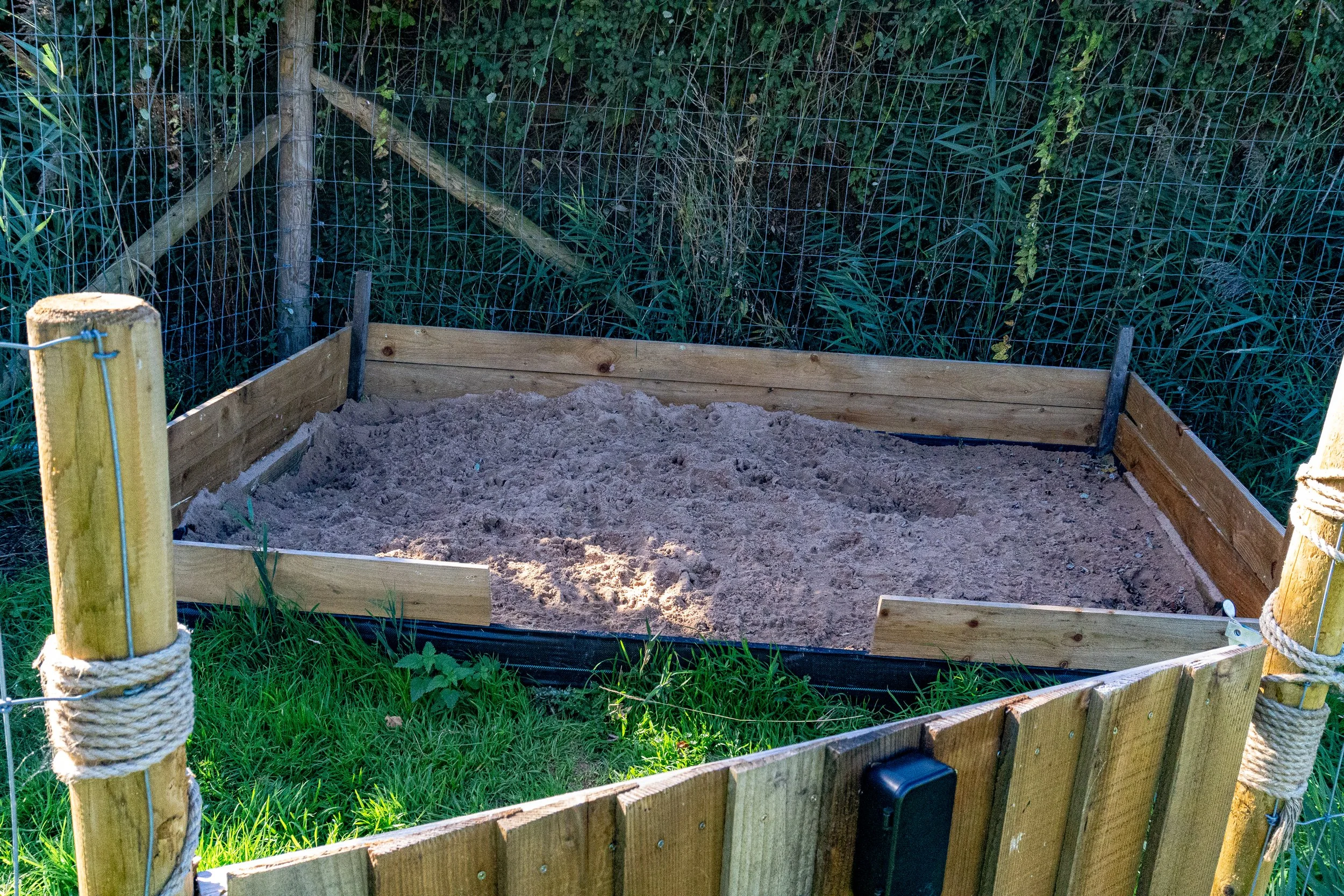 A small fenced enclosure with sand at the bottom, surrounded by a wire and wooden fence with rope ties, next to tall grass and green vegetation.