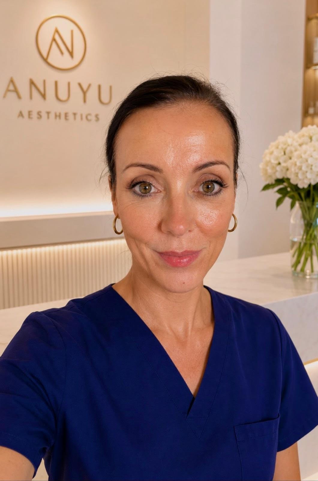 A woman in a blue medical uniform taking a selfie inside a clinic or aesthetic center with a sign that says "ANUYU AESTHETICS" on the wall behind her, alongside a vase of white flowers.