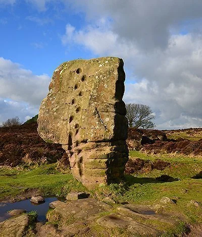Large ancient standing stone in a grassy field with a small pond nearby, under a partly cloudy sky.