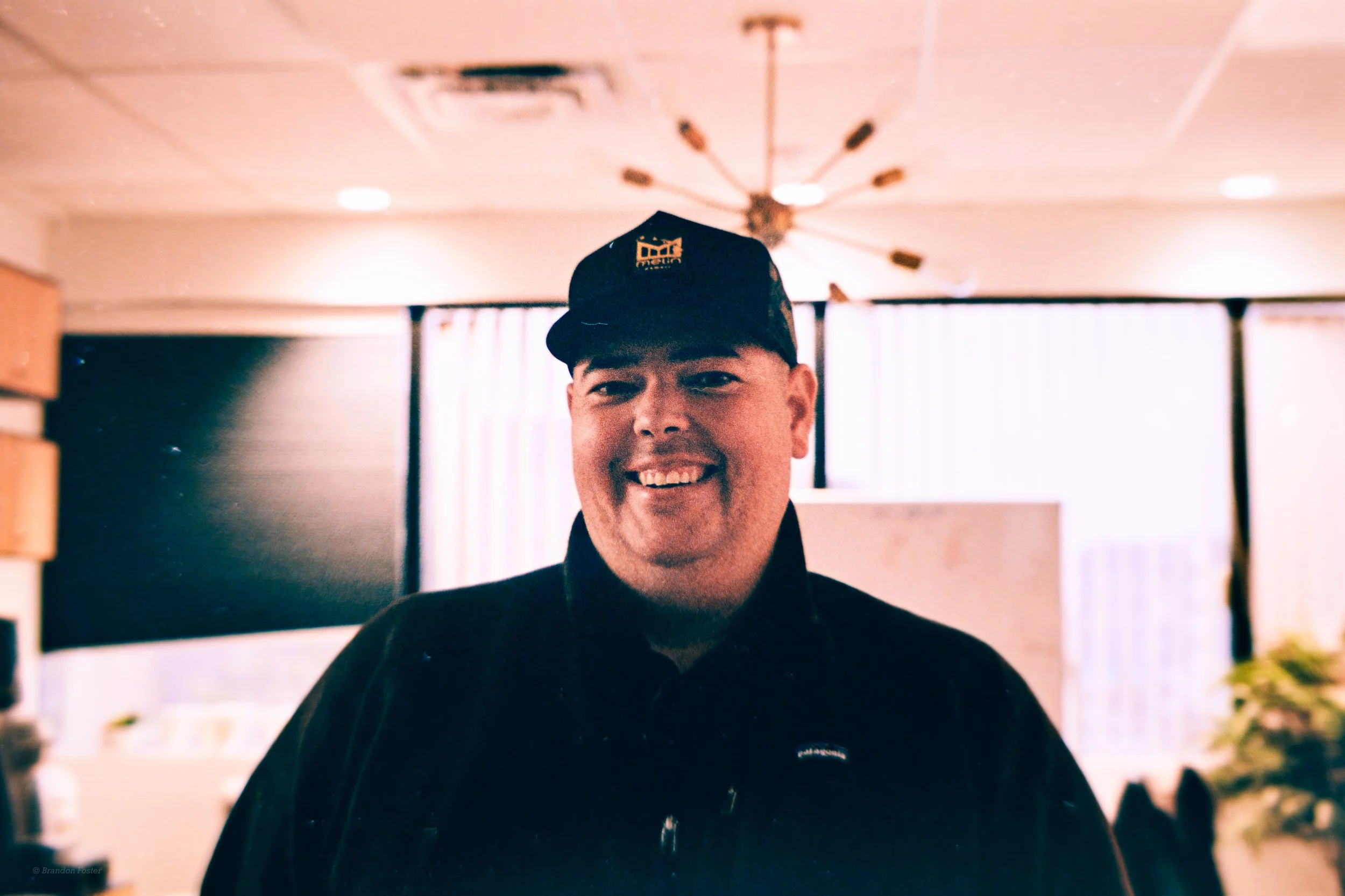 A young man smiling, wearing a black cap and black jacket, standing indoors with a modern chandelier in the background.