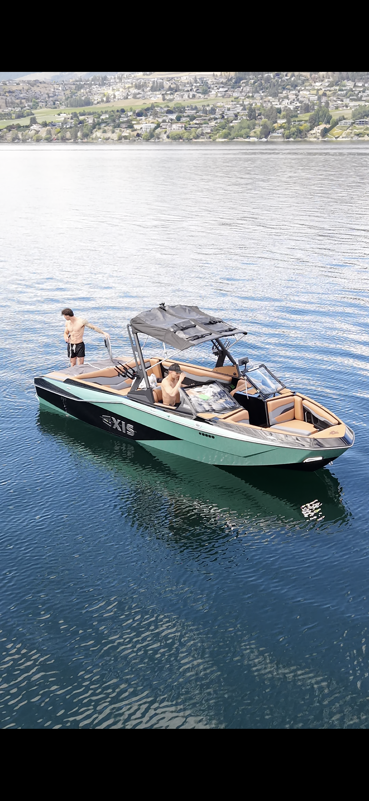 Two shirtless men are on a green and black boat with tan interior, one sitting at the controls and the other standing at the rear, on a calm body of water with a hilly town in the background.