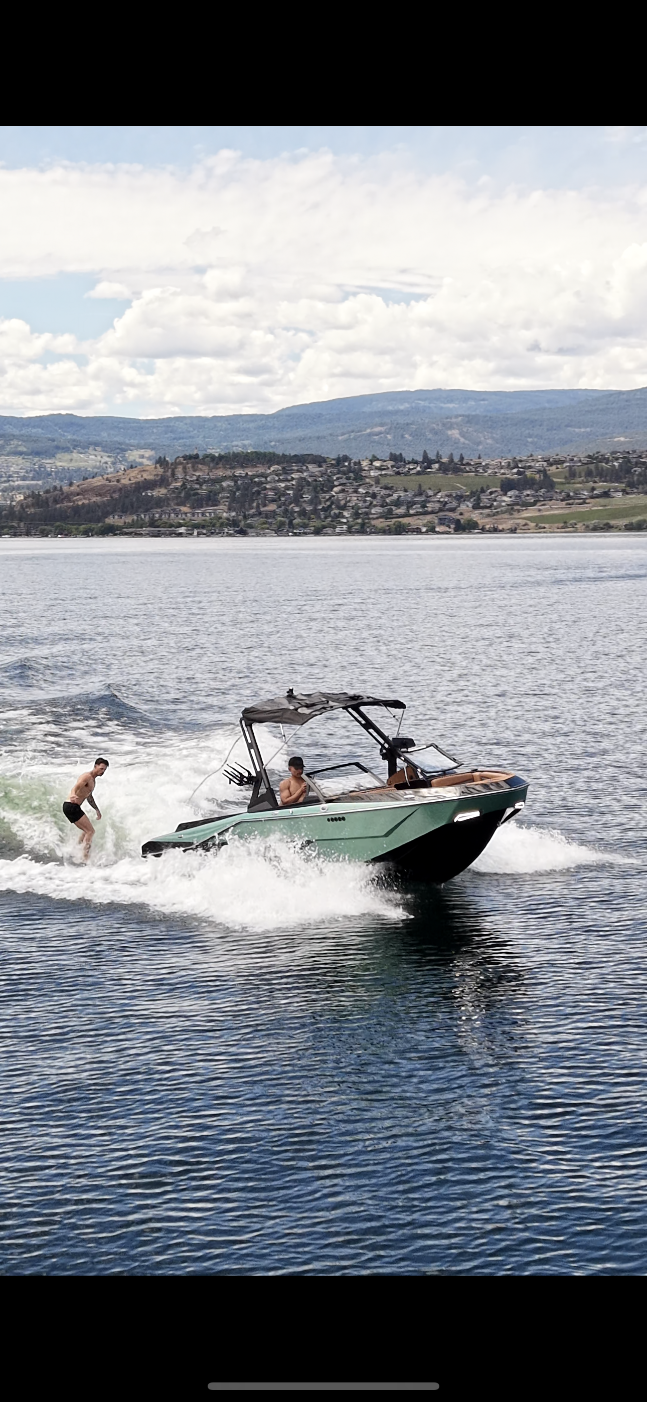 A boat speeds across the water with two young men on board, one sitting and another standing at the rear, with a third young man jumping into the water nearby. The background shows a scenic landscape with hills and a partly cloudy sky.