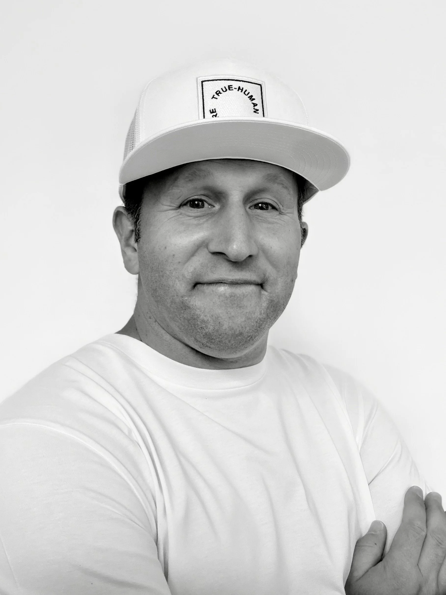 A man wearing a white cap with the text 'TRUE-HUMAN' on it, and a white shirt, standing against a plain background.