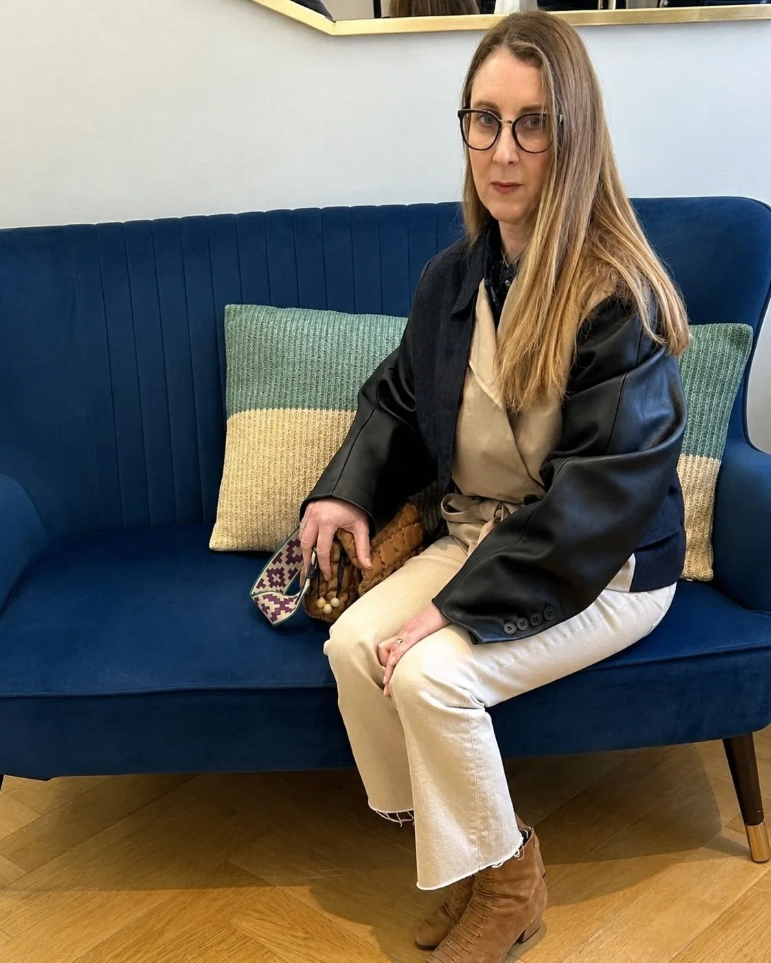Sam Remer sitting on a blue sofa wearing a black jacket and cream jeans