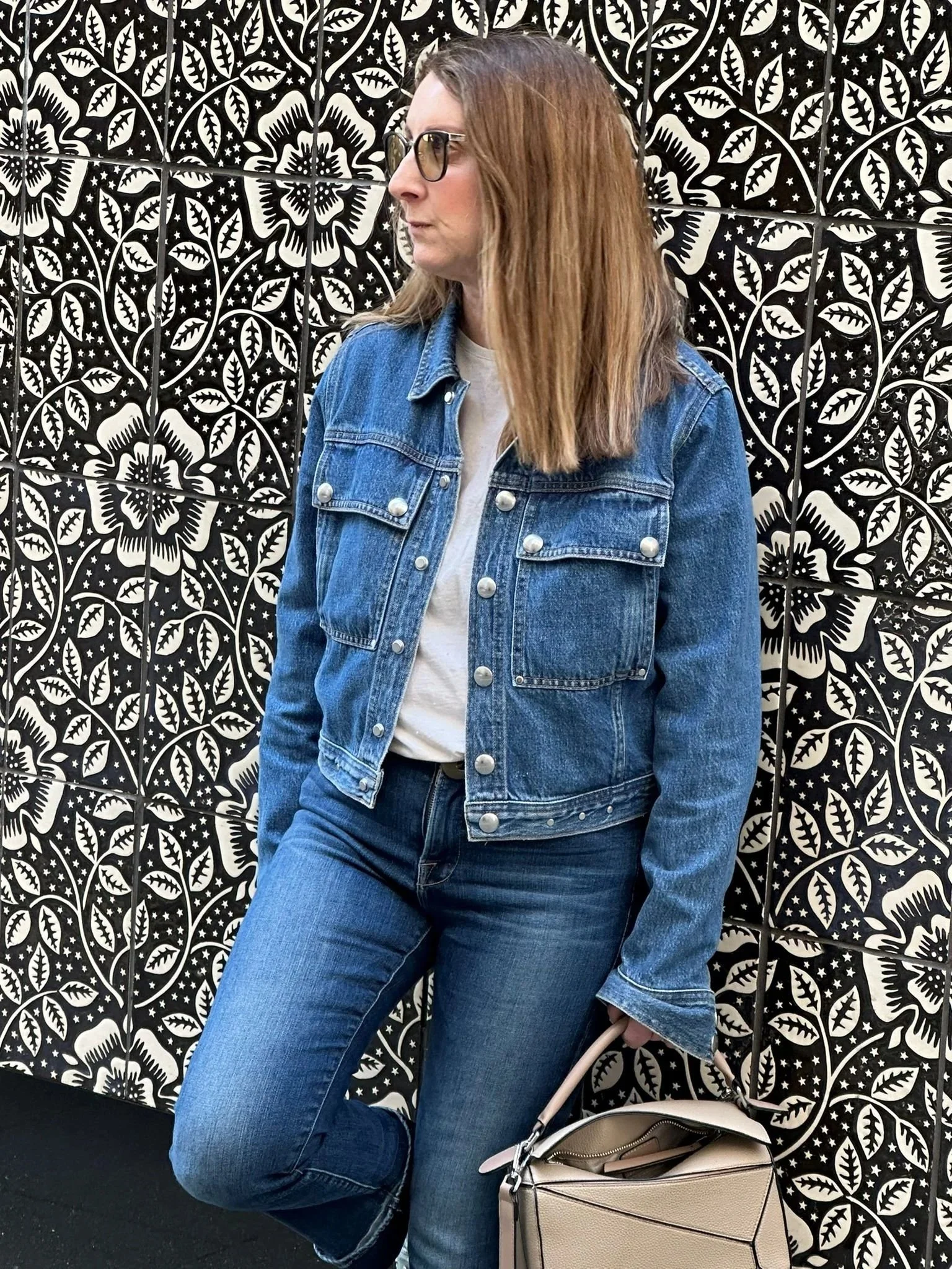 Sam Remer wearing a denim jacket and light top, standing against a black and white floral backdrop.