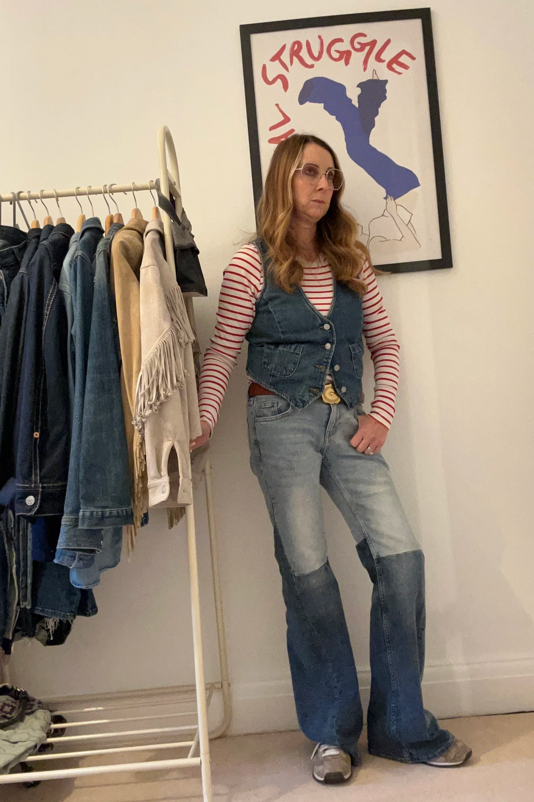 A woman with long wavy hair, wearing glasses, a red and white striped shirt, a denim vest, and baggy jeans, stands indoors next to a clothing rack holding various denim and casual wear. Behind her is a framed poster with the words "My Struggle" and an illustration of a reclining figure.