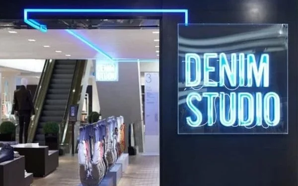 Neon sign reading 'Denim Studio' at Selfridges denim studio entrance with escalators in the background.