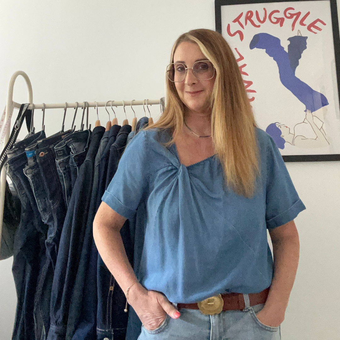 A woman with long blonde hair, glasses, and a blue top standing in front of a clothing rack with jeans and pants, and a wall poster with the words 'Struggle' and a figure of a woman.