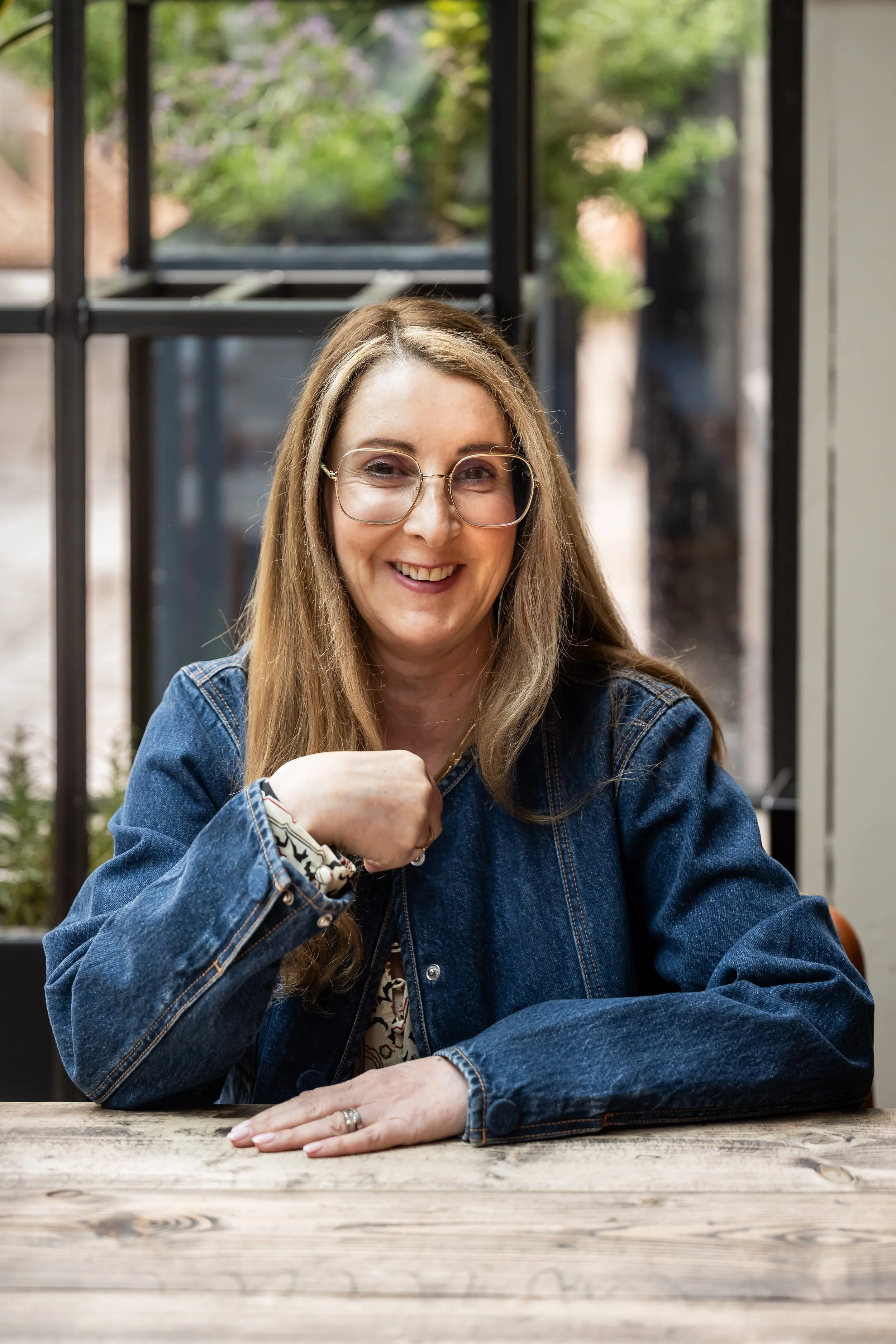 A smiling woman with glasses and long hair, wearing a denim jacket, sitting at a rustic wooden table indoors with a background of large windows and greenery outside.
