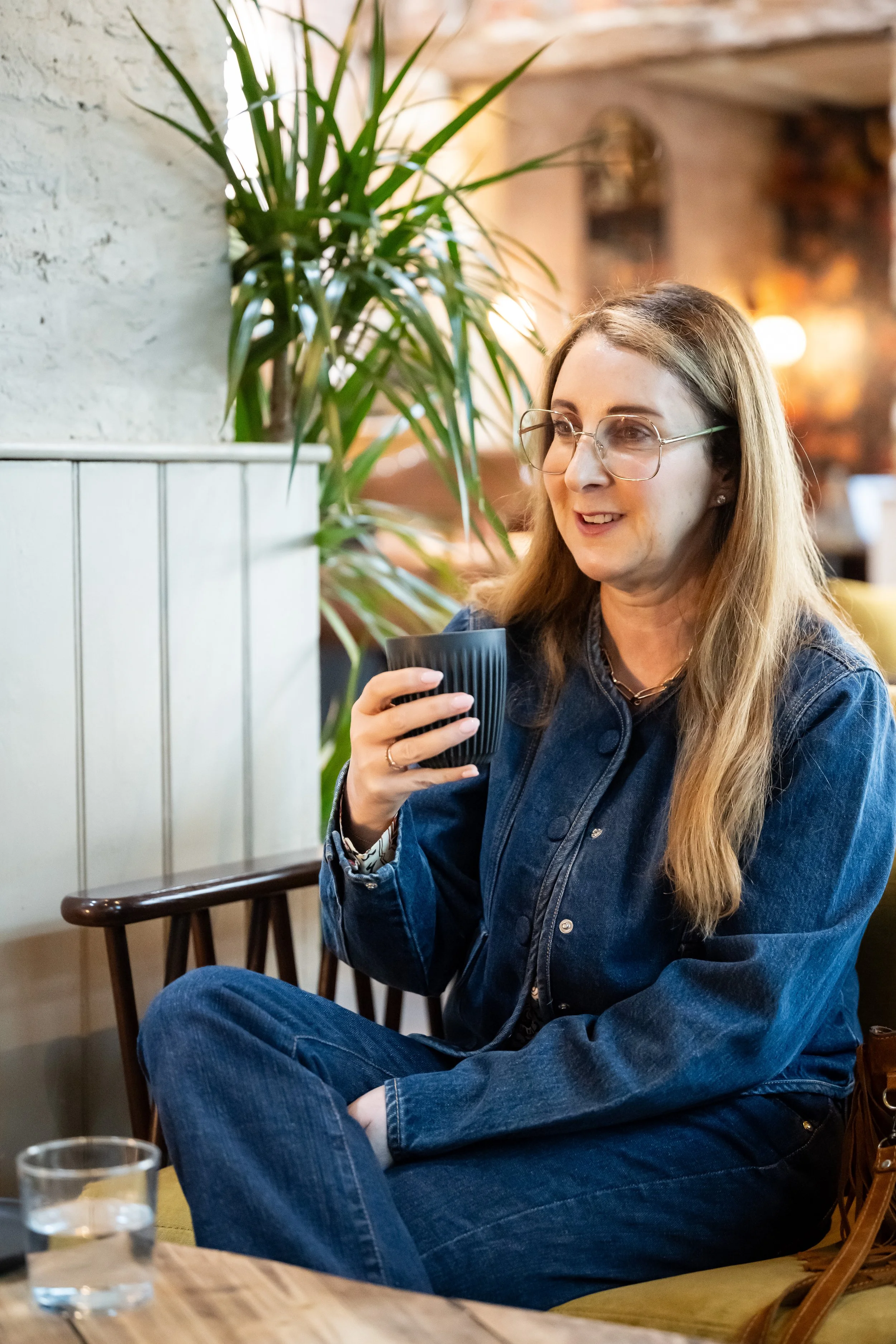 A woman with long blond hair, glasses, and a denim jacket sitting in a cozy cafe, holding a dark cup, with a large green plant behind her and warm lighting in the background.