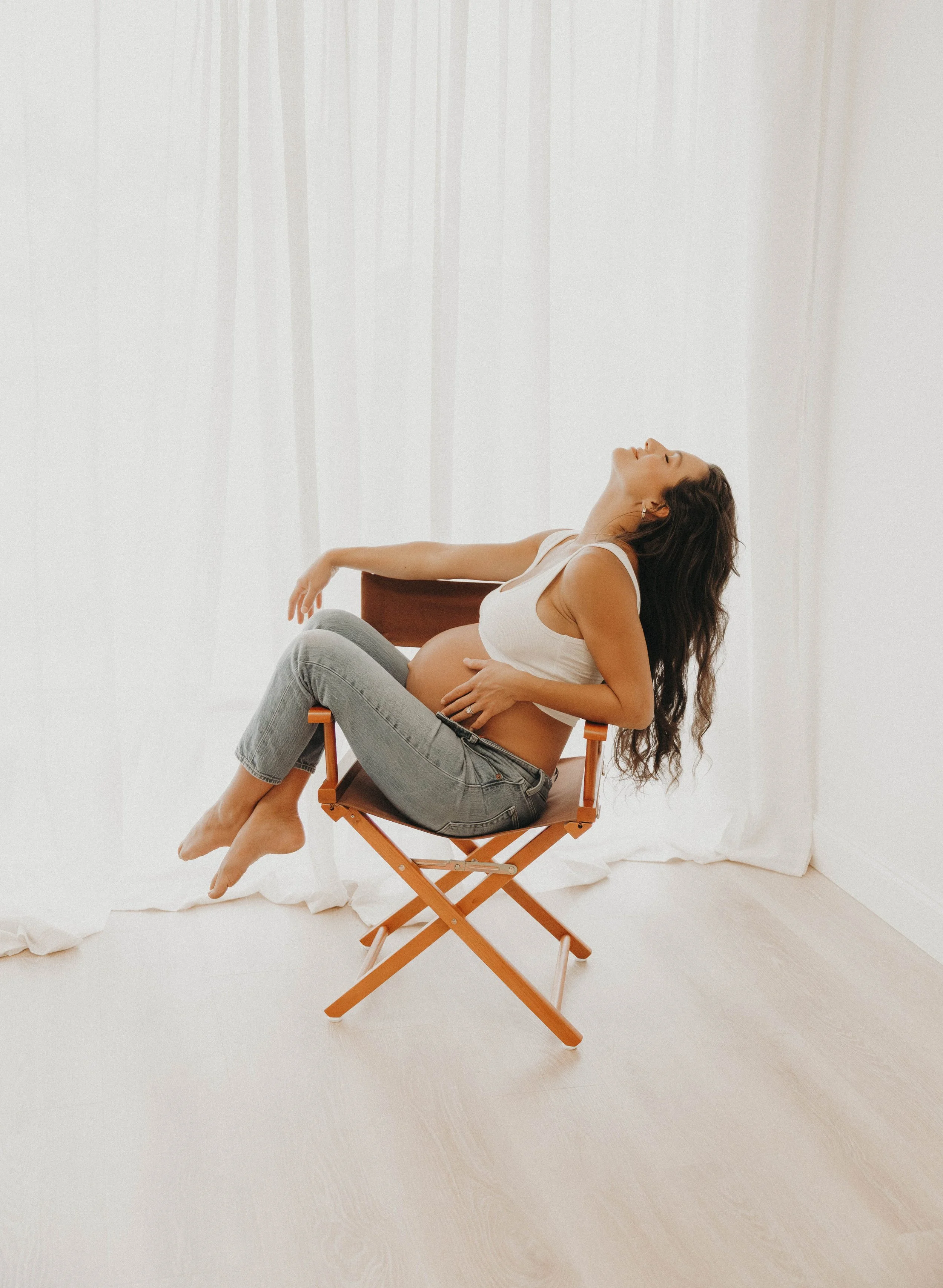 A pregnant woman sitting on a wooden folding chair in a minimalist white room, with her eyes closed and head tilted back, resting her hand on her belly.