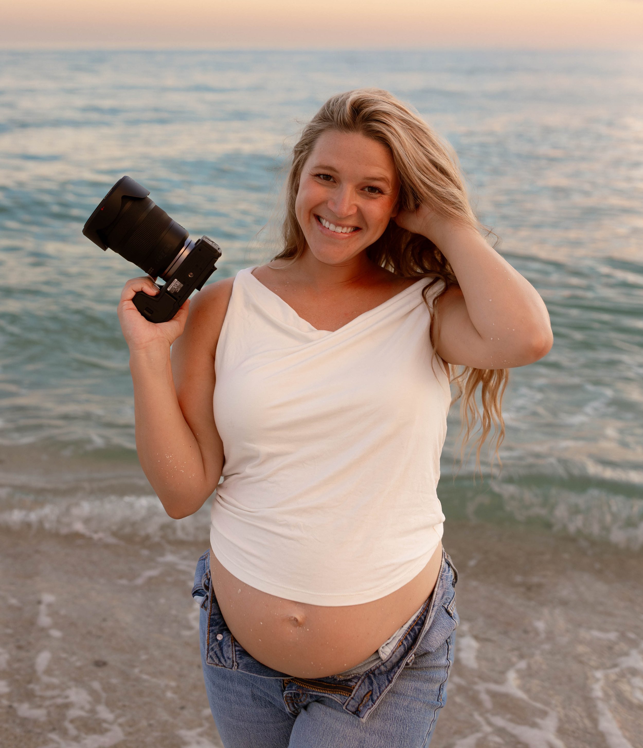 Young woman smiling at the camera on a beach, holding a camera in her right hand, wearing a white sleeveless top and denim shorts, with the ocean and sunset in the background.