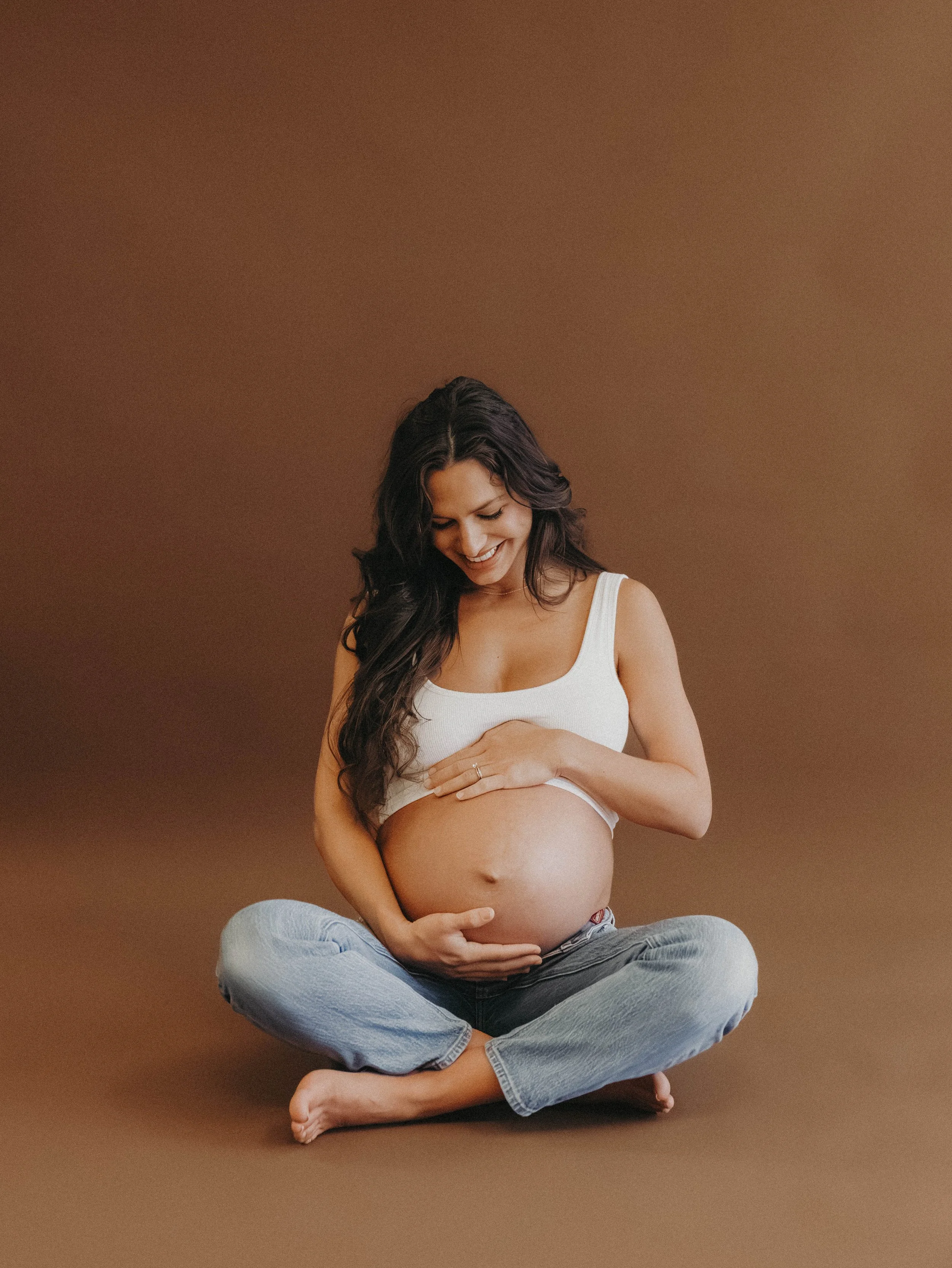 A pregnant woman sitting cross-legged on the floor, smiling and looking down at her baby bump, against a brown background.