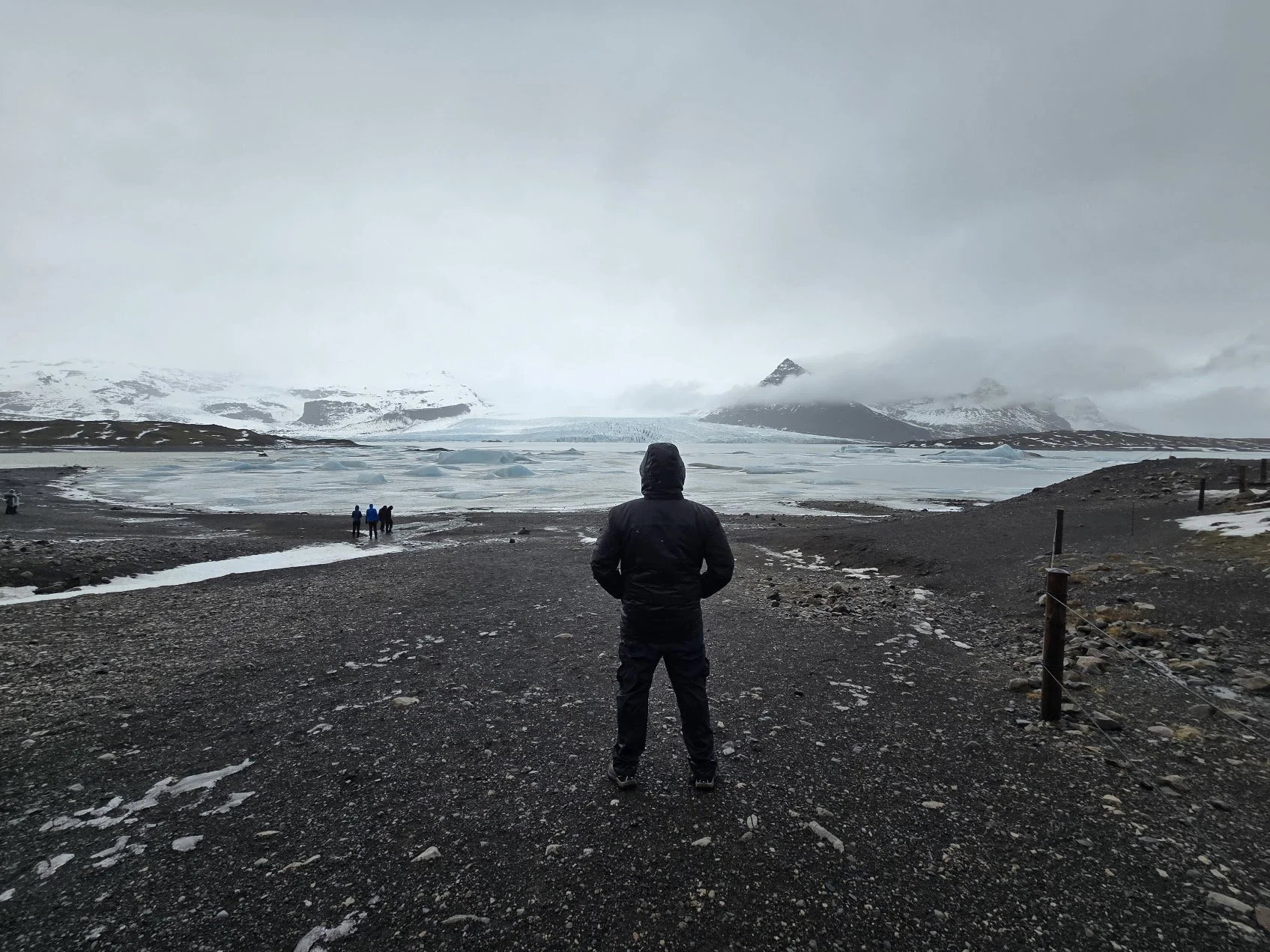 Person in black jacket with hood standing on rocky beach, overlooking icy, snow-covered landscape with distant mountains, glaciers, and a cloudy sky.