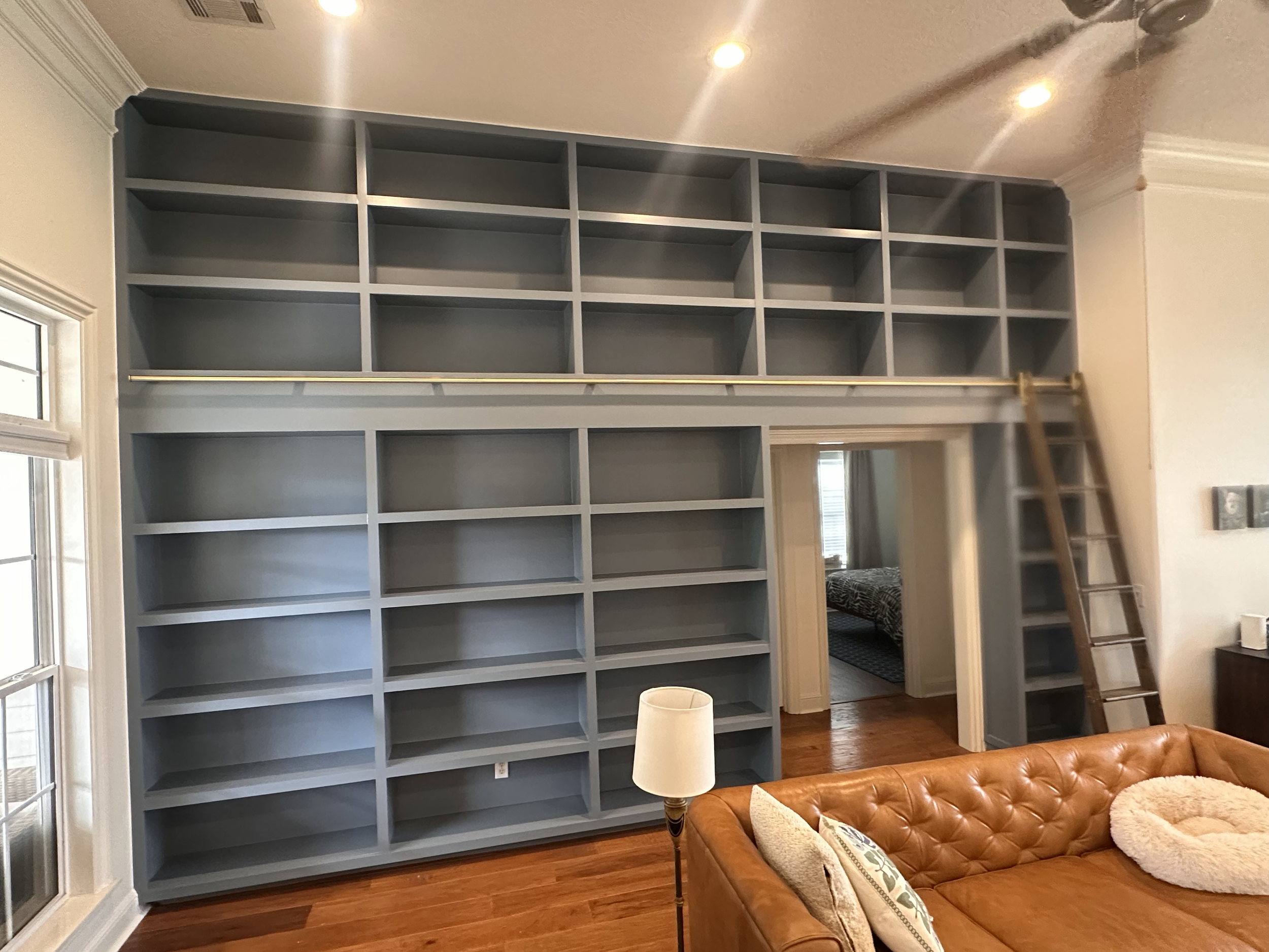Empty blue bookshelf in a living room with a large window, a lamp, a leather couch with pillows, and a wooden ladder.