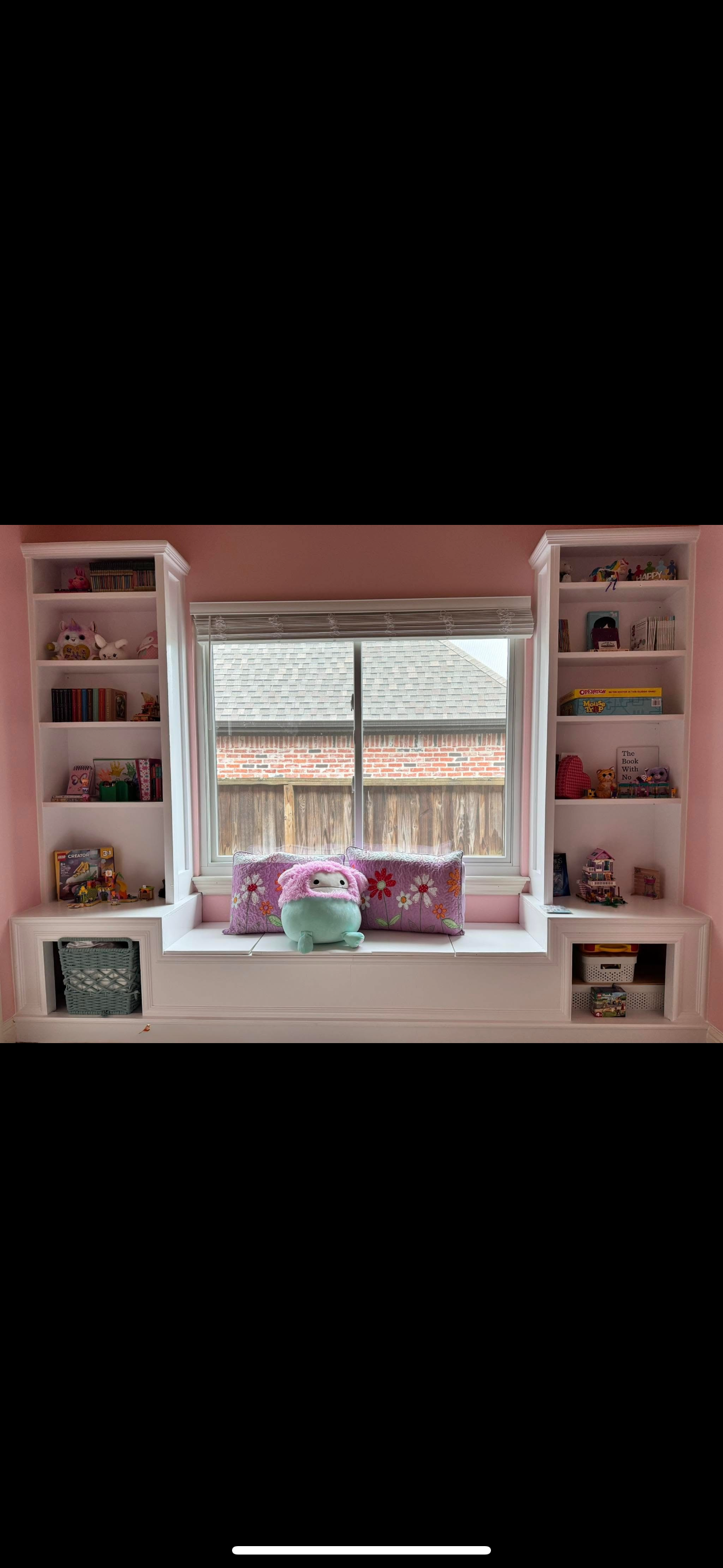 A window seat with pink floral cushions and a plush toy in front of a window in a pink-walled room. White bookshelves with toys and books flank the window.