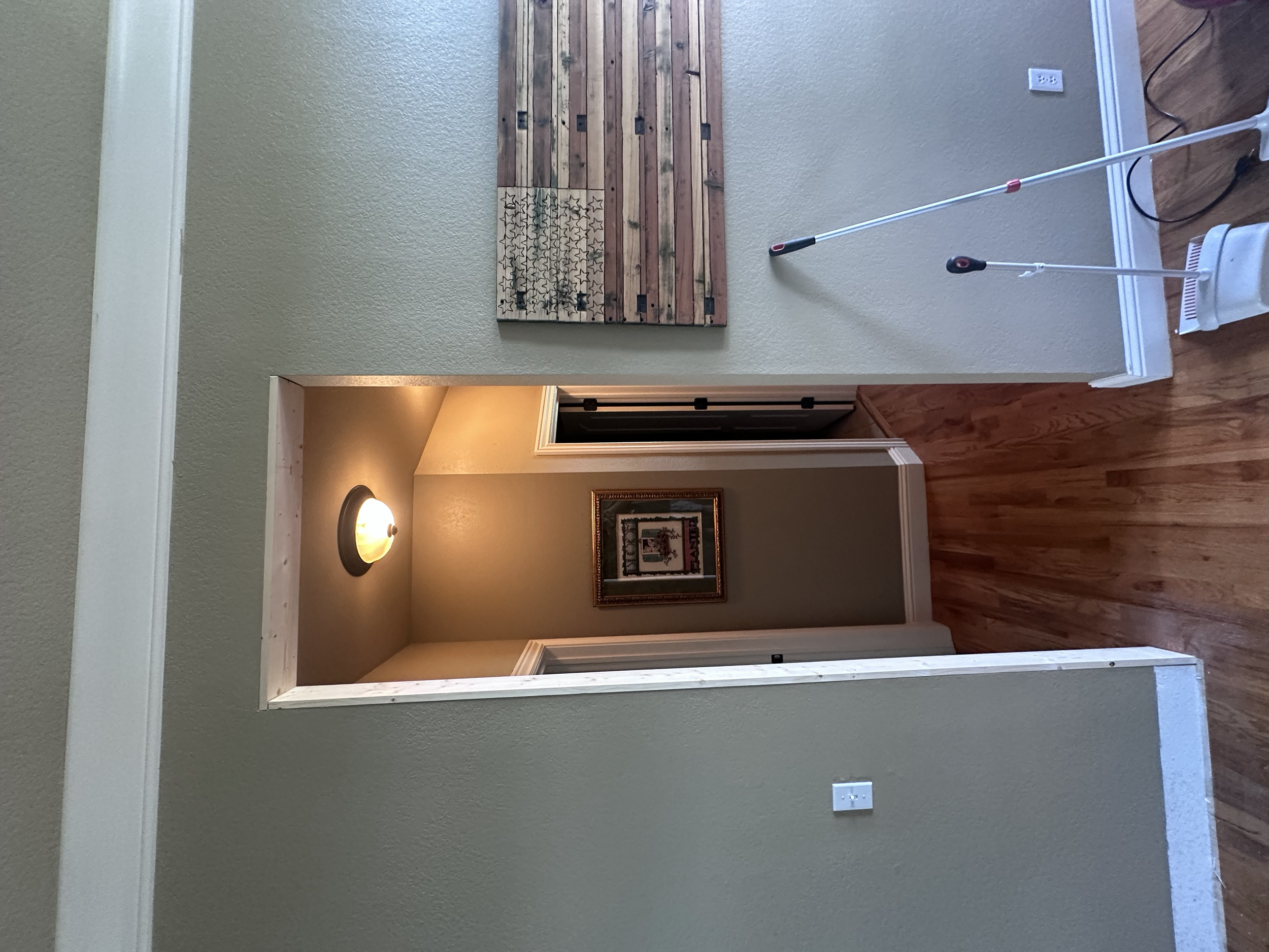 A hallway with beige walls and wooden flooring. On the wall, there is a decorative firewood holder made of wooden planks, and below it, a star-shaped metal decoration. To the right, there is an electric outlet and a white portable heater with a black