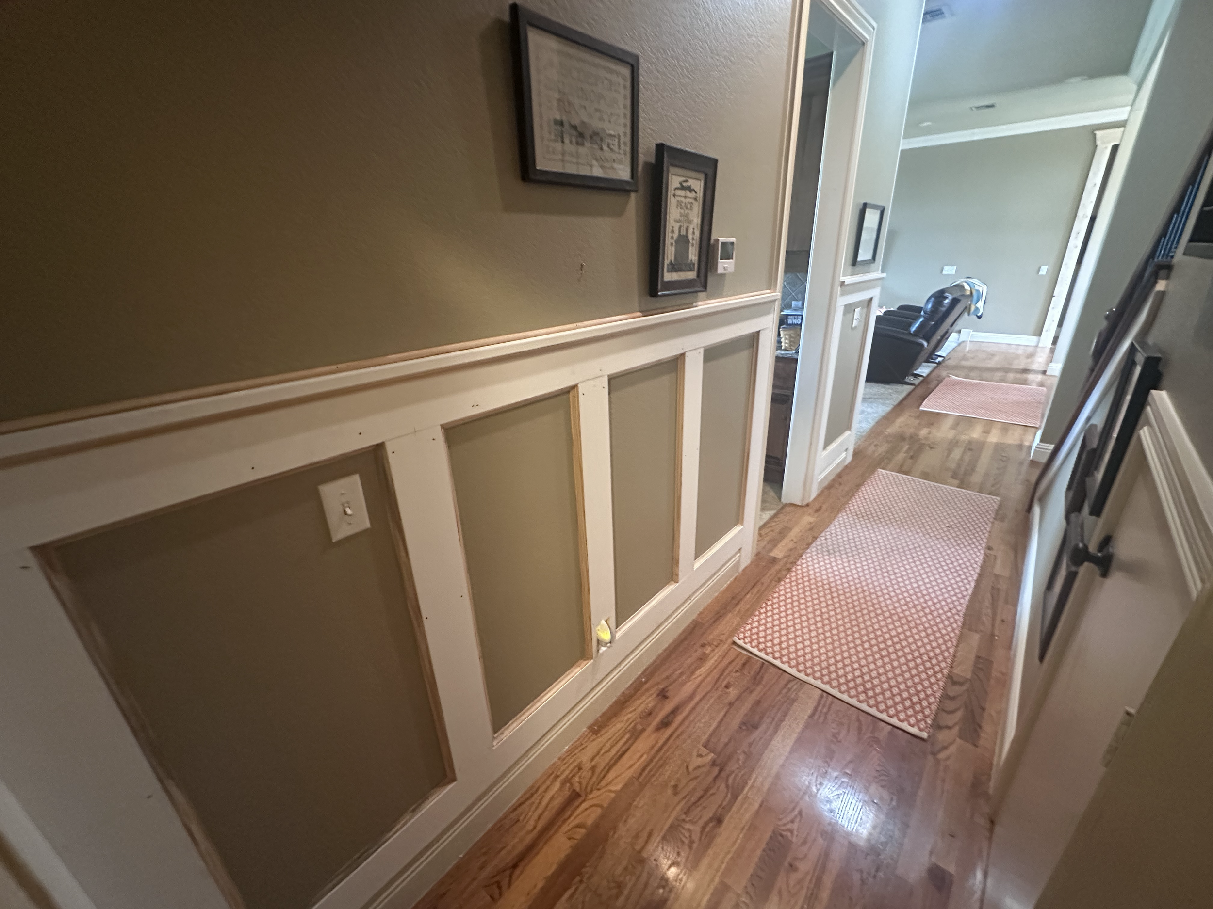 Hallway with hardwood flooring, pink rug, framed pictures on the wall, and a seating area visible in the background.