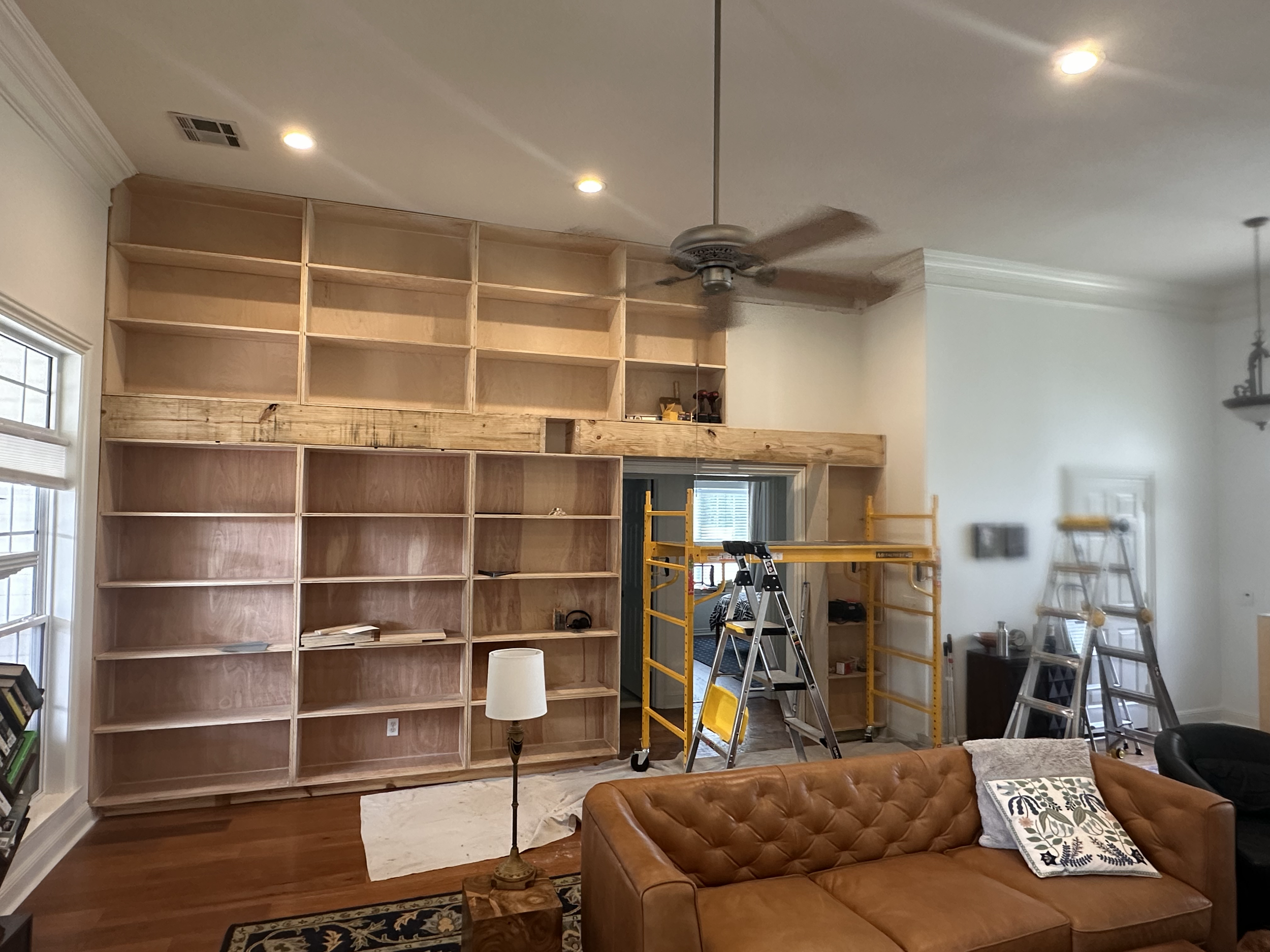 Living room under construction with empty wooden bookshelves, a ladder, a scaffolding, a standing lamp, a ceiling fan, and a brown leather sofa with a patterned pillow.