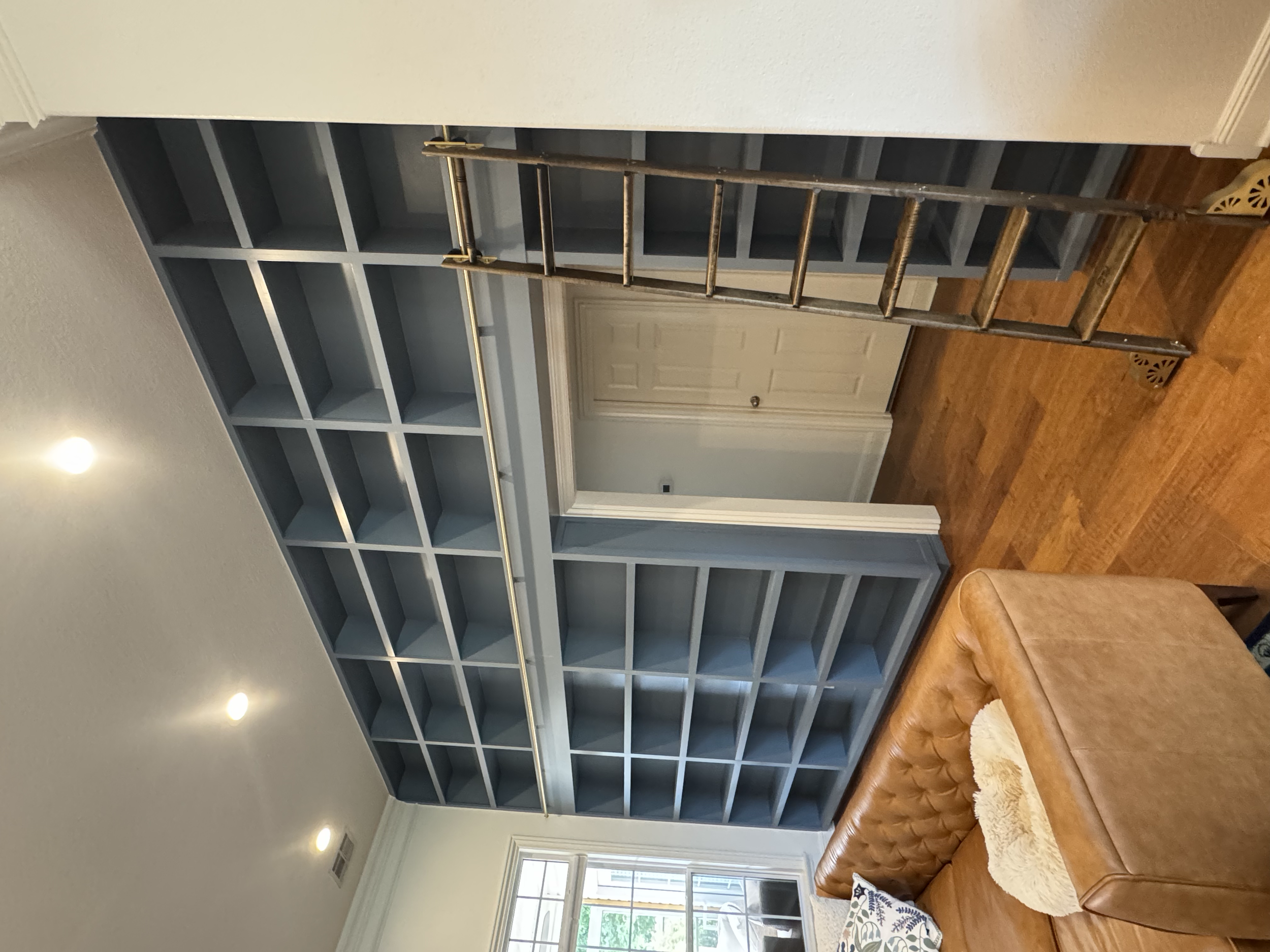 Empty built-in bookshelf with multiple shelves, a ladder, and a couch with decorative pillows in a living room with hardwood floors and a large window.