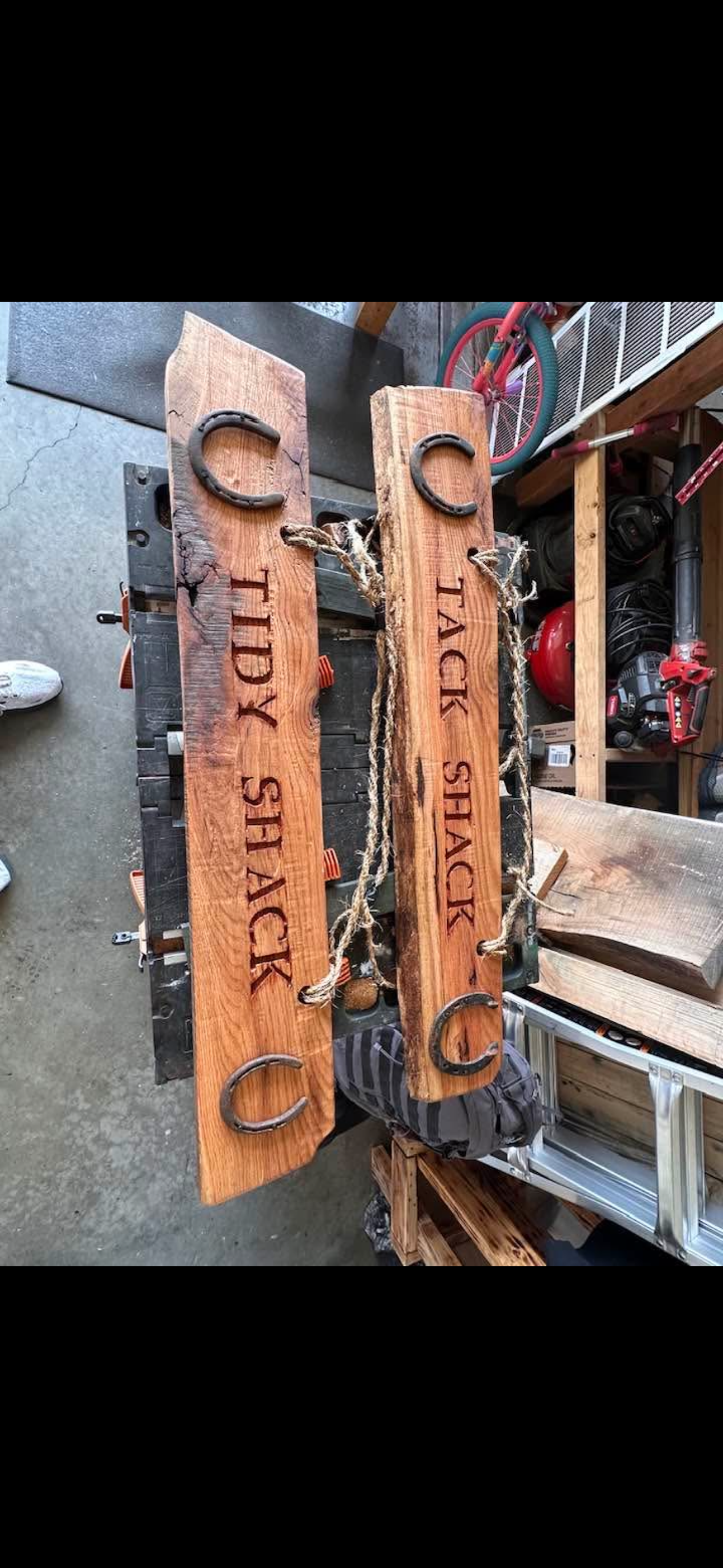 Two wooden signs with the words 'CACK IDY STAY' and 'CACK STAY' carved into them, decorated with horseshoes and ropes, resting on a workbench in a workshop.