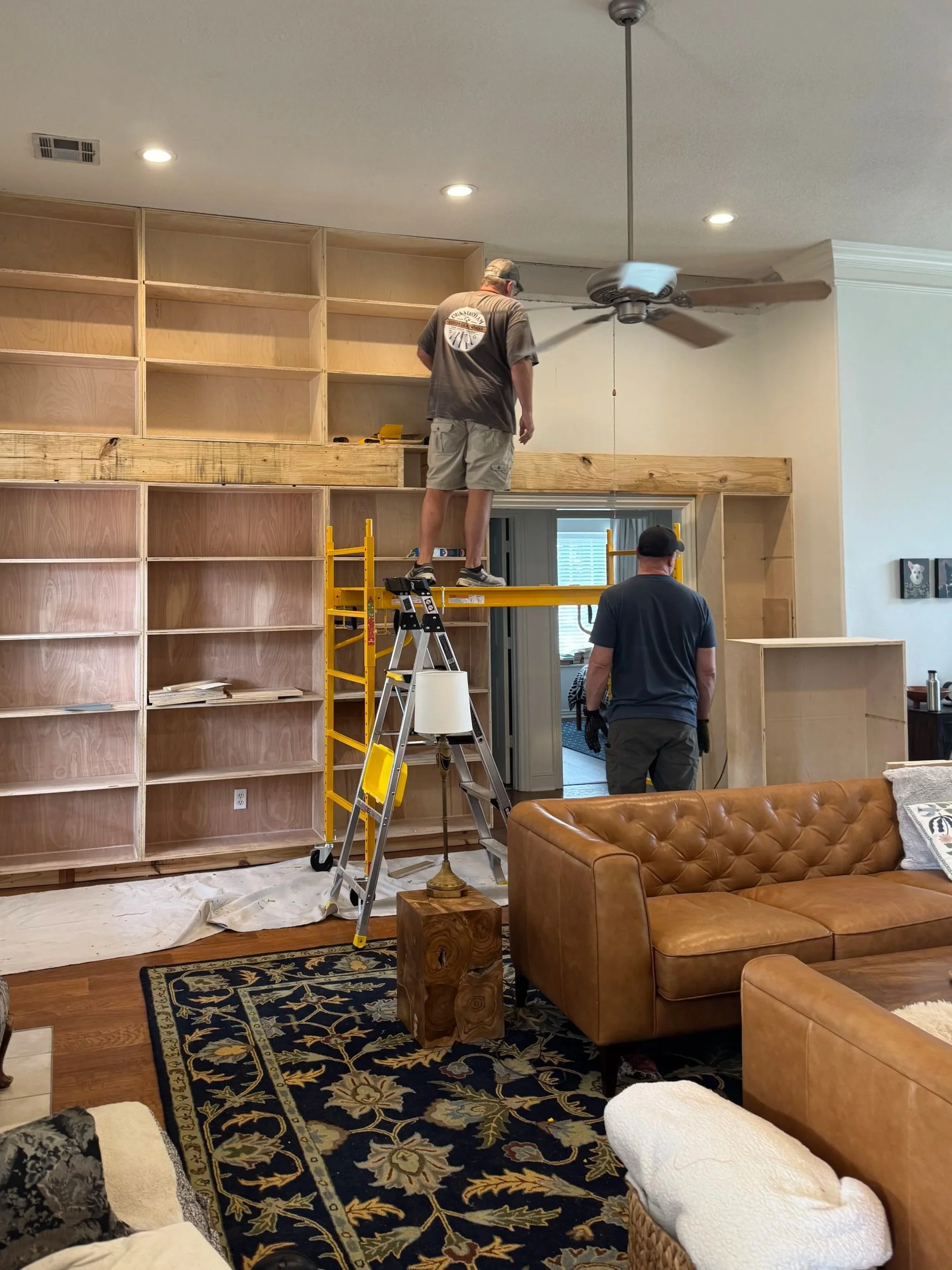 Two men are installing wooden shelving and a wood beam in a living room with a high ceiling. One man is standing on a yellow step ladder, working near the ceiling fan, while the other stands below, supervising. The room has a large, detailed dark area rug, a tan leather couch, and a basket with white blankets and pillows.