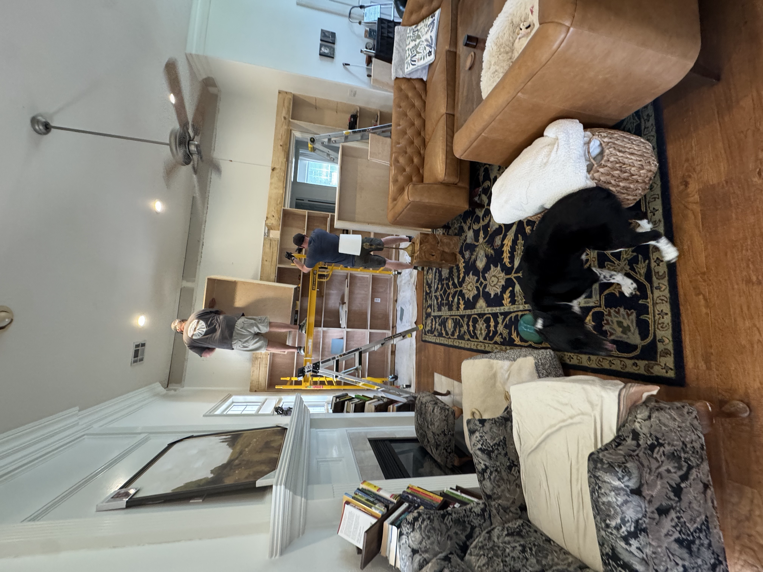People working on furniture assembly in a living room, using a ladder and tools, surrounded by bookshelves, a carpet, and a leather couch.