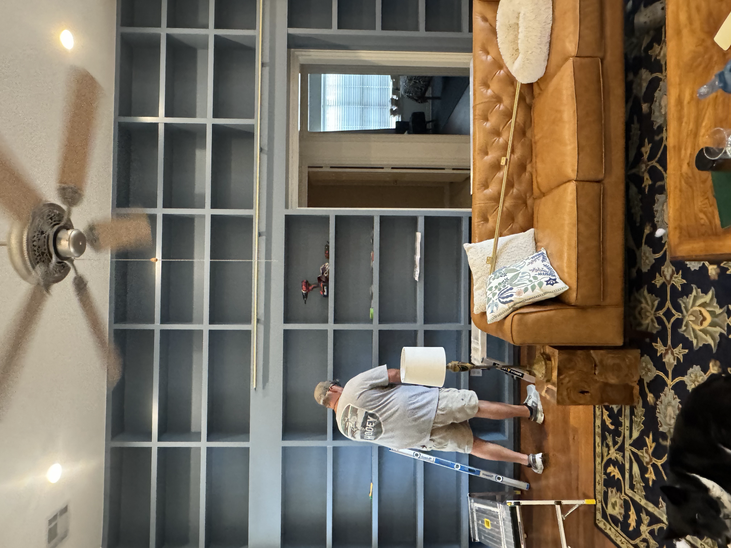 A man in a white shirt is installing a large gray shelving unit in a living room. The room has a leather sofa, a wooden coffee table, a decorative rug, and a man sitting on the floor. There are windows with blinds in the background.