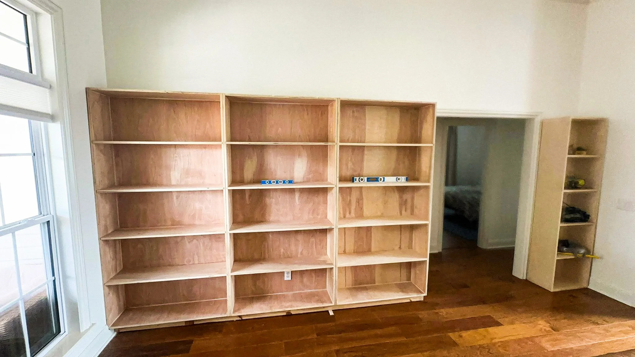 Unfinished wooden bookshelf against a white wall near a doorway, with a pair of blue spirit levels on it, in a room with hardwood flooring and a sliding glass door on the left.