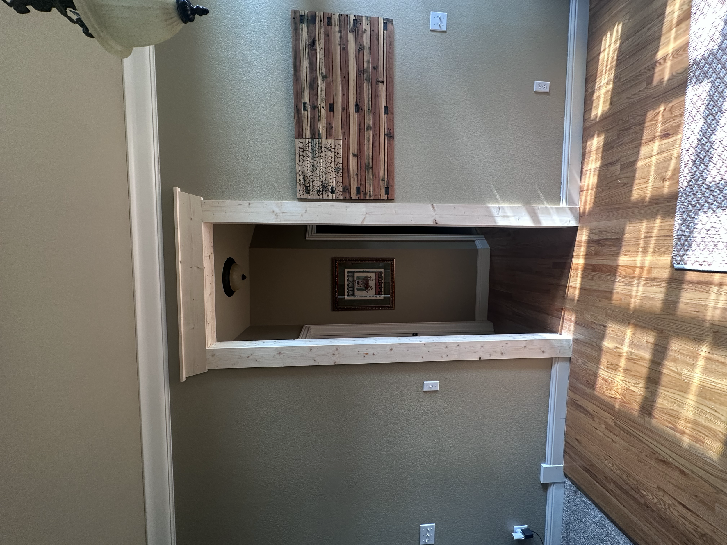 Interior of a room with a green wall, a wooden frame structure with hinges, a framed picture, and a wooden floor with sunlight casting shadows.