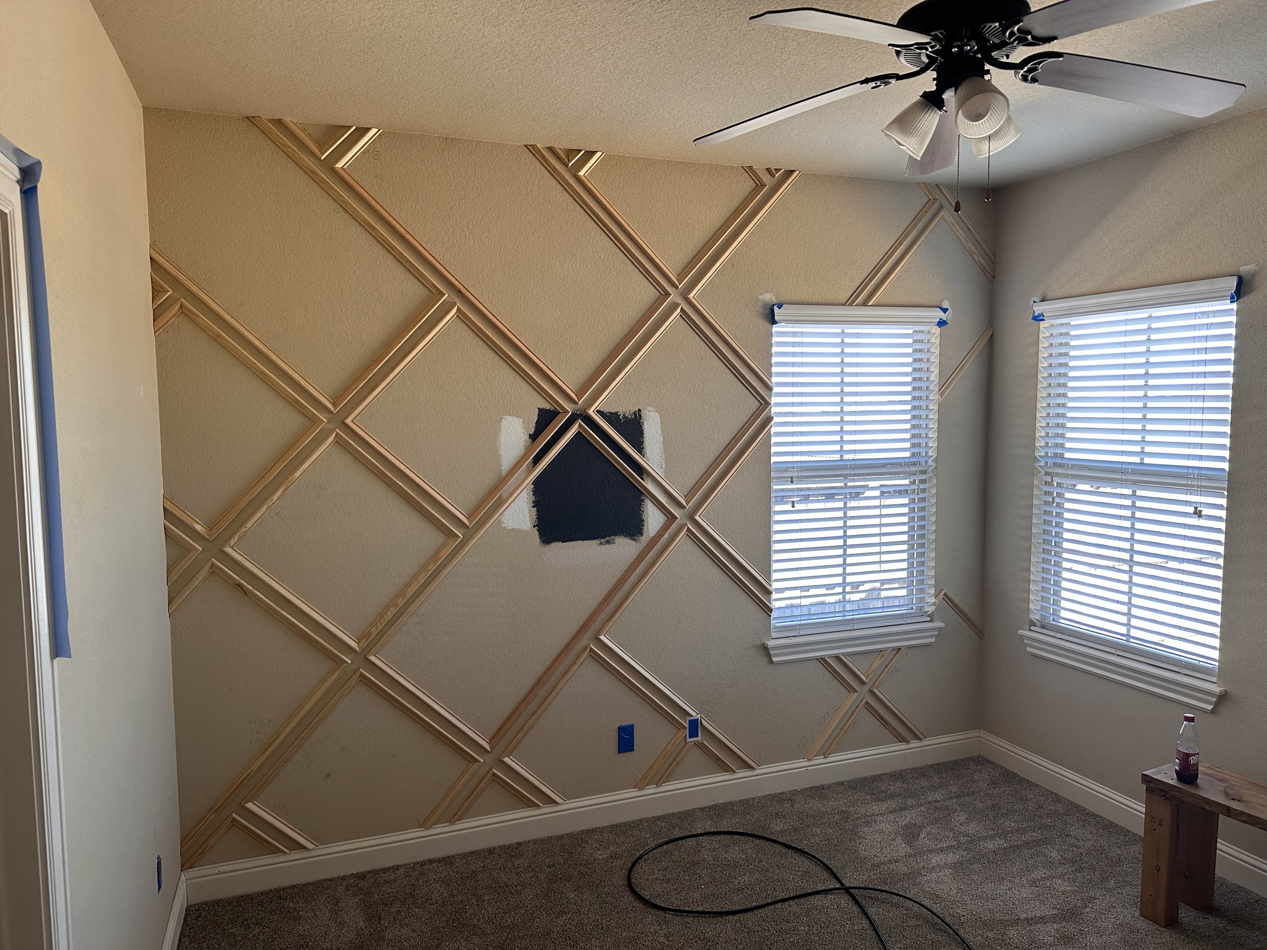 Interior of a room with beige walls, two windows with white blinds, and a ceiling fan. One wall has an intricate diamond-shaped wood frame pattern, with an area painted dark blue. There are paint supplies and a wooden stand on the floor.