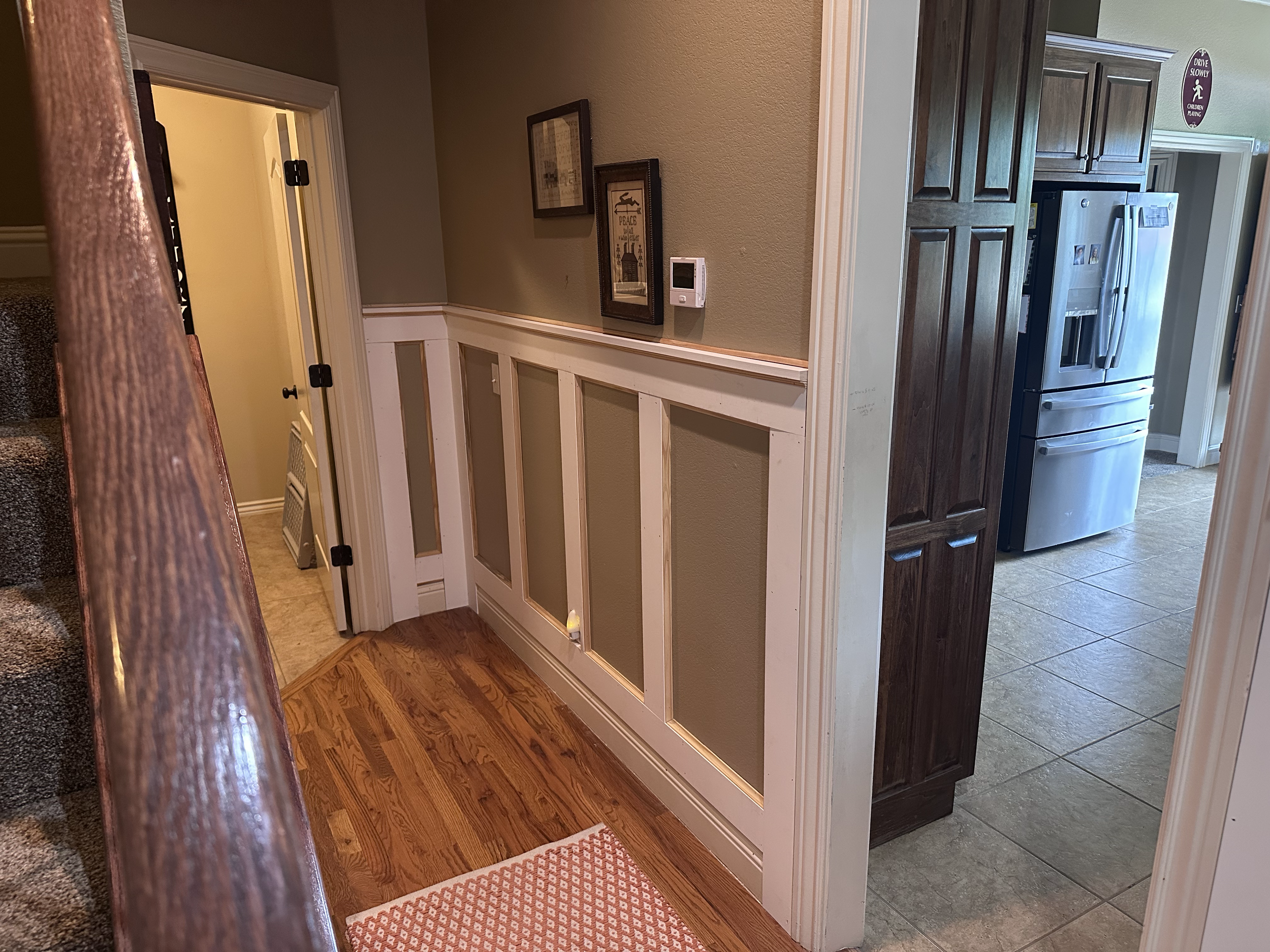 Interior view of a kitchen with wooden floors, a beige wall, and an unfinished half-wall with white trim. A framed picture hangs on the wall. There is a stainless steel refrigerator and dark wood cabinets. Part of a staircase with a wooden handrail i