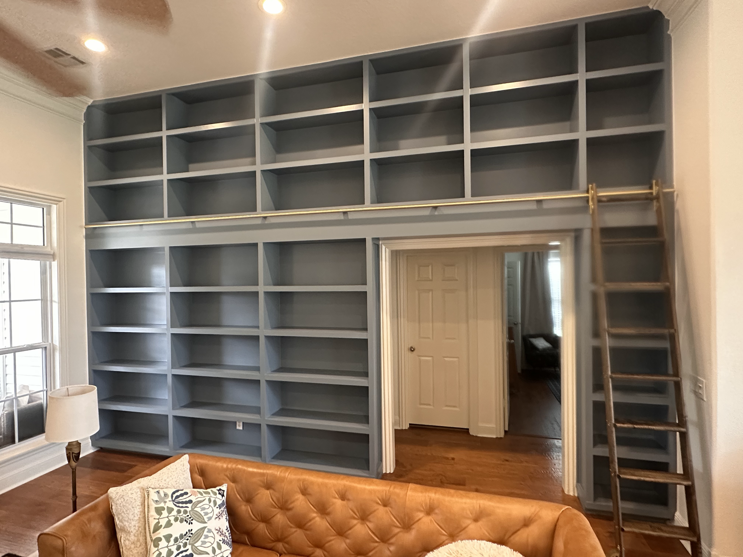A living room with a large built-in gray bookshelf and a ladder on the right side. A brown leather tufted couch with patterned pillows is in the foreground. A window is on the left and a door is visible in the background.
