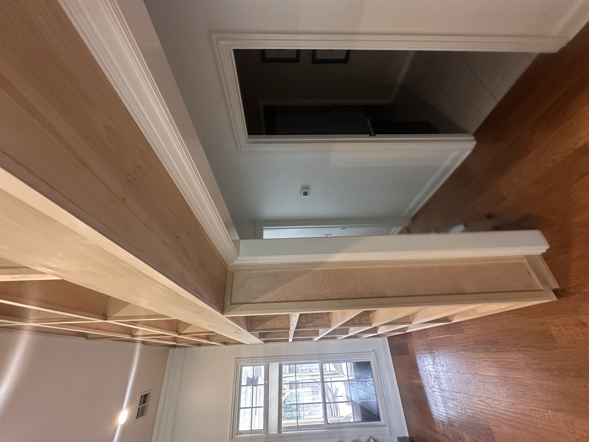 Interior view of a residential space with wooden flooring, light-colored walls, and a large window allowing natural light. A doorway leads to another room, and framed pictures are hung on the wall. The photo is rotated 90 degrees clockwise.