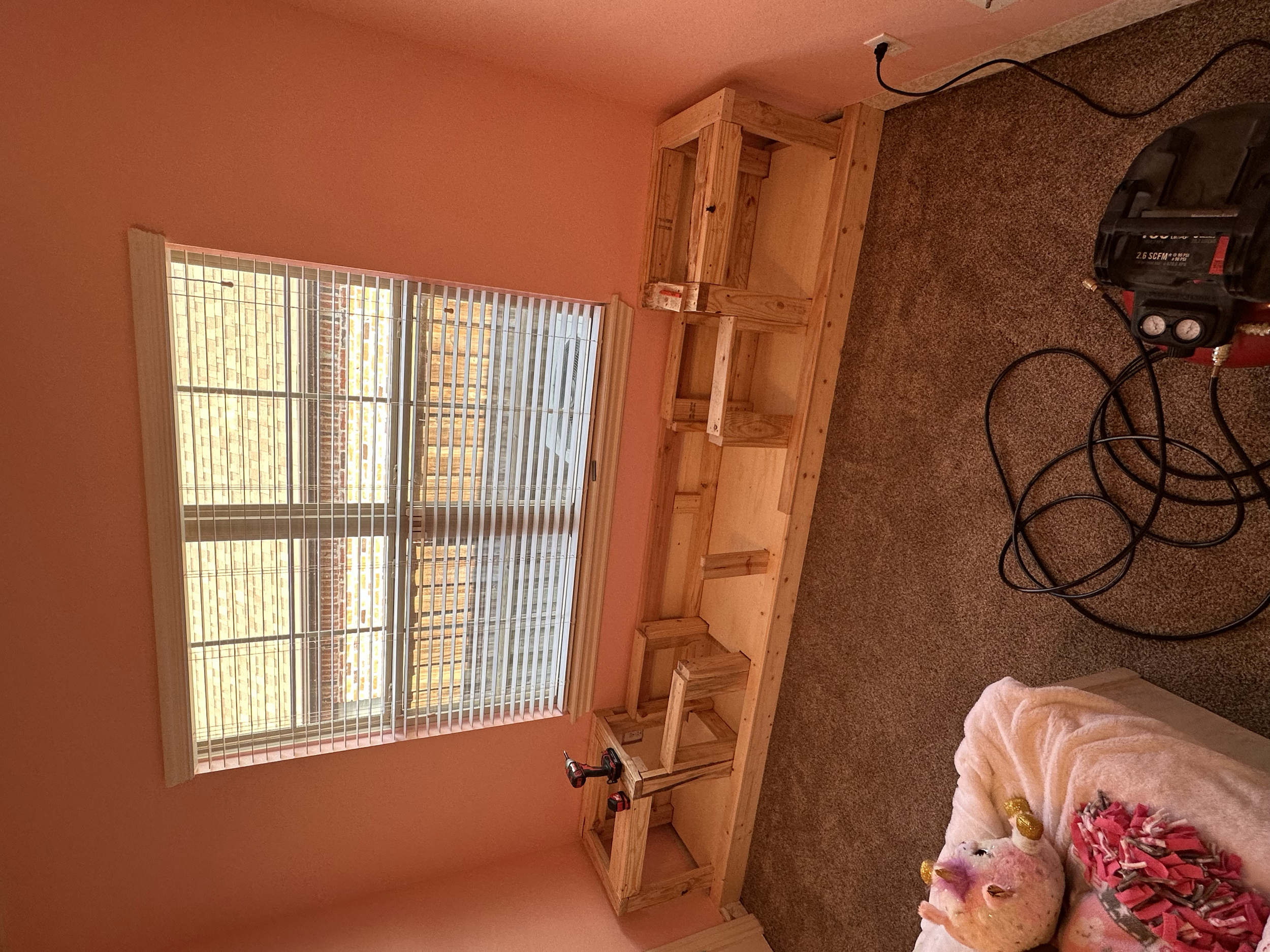 A room under renovation with an unfinished wooden bookshelf, a window with blinds, and a carpeted floor. There is a cordless drill on the floor and some stuffed animals and toys in a basket.