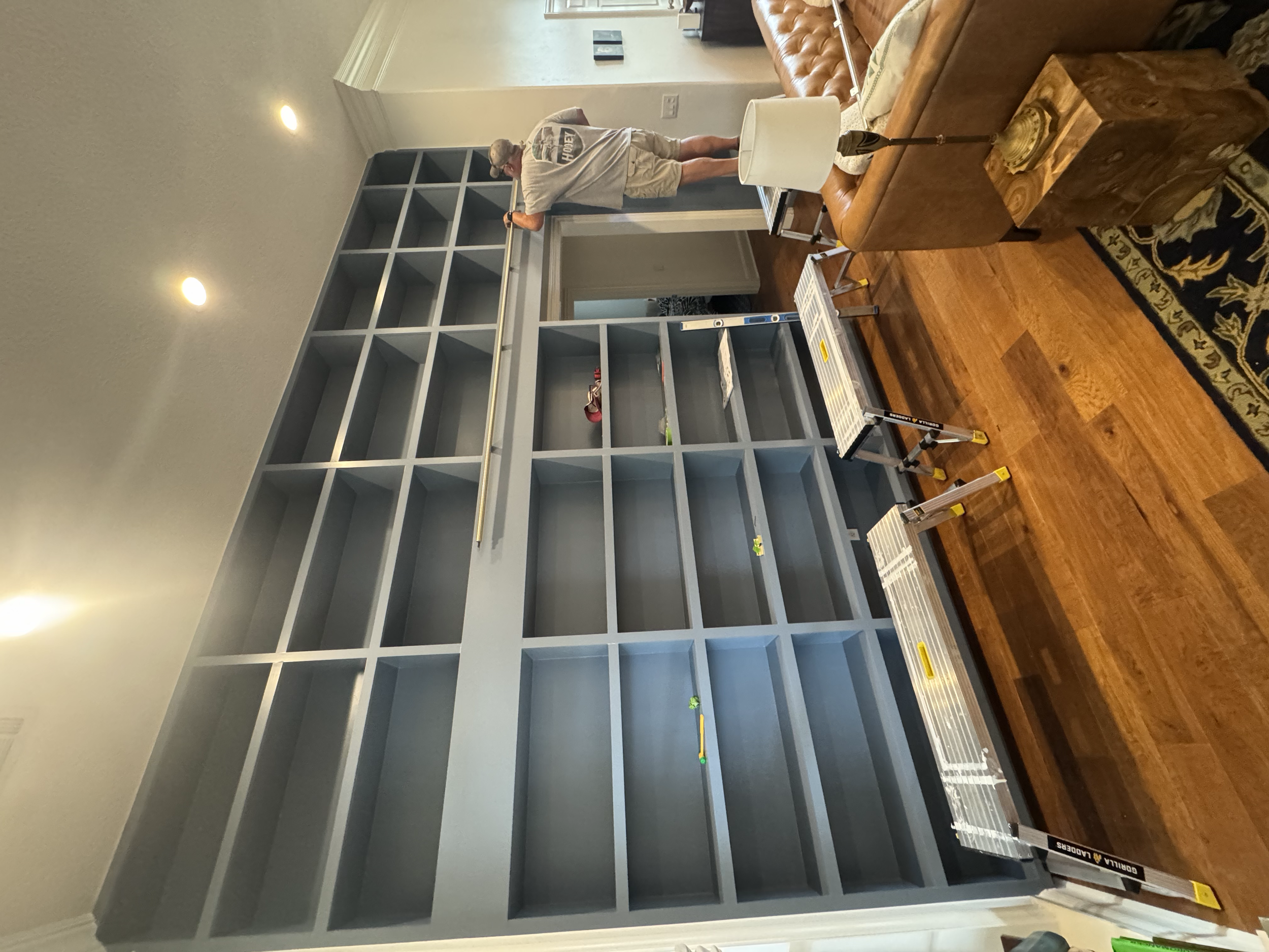 An empty gray shelving unit is being assembled in a living room by a man using a step ladder. There is a brown leather sofa, a side table, a lamp, and a rug on the hardwood floor.