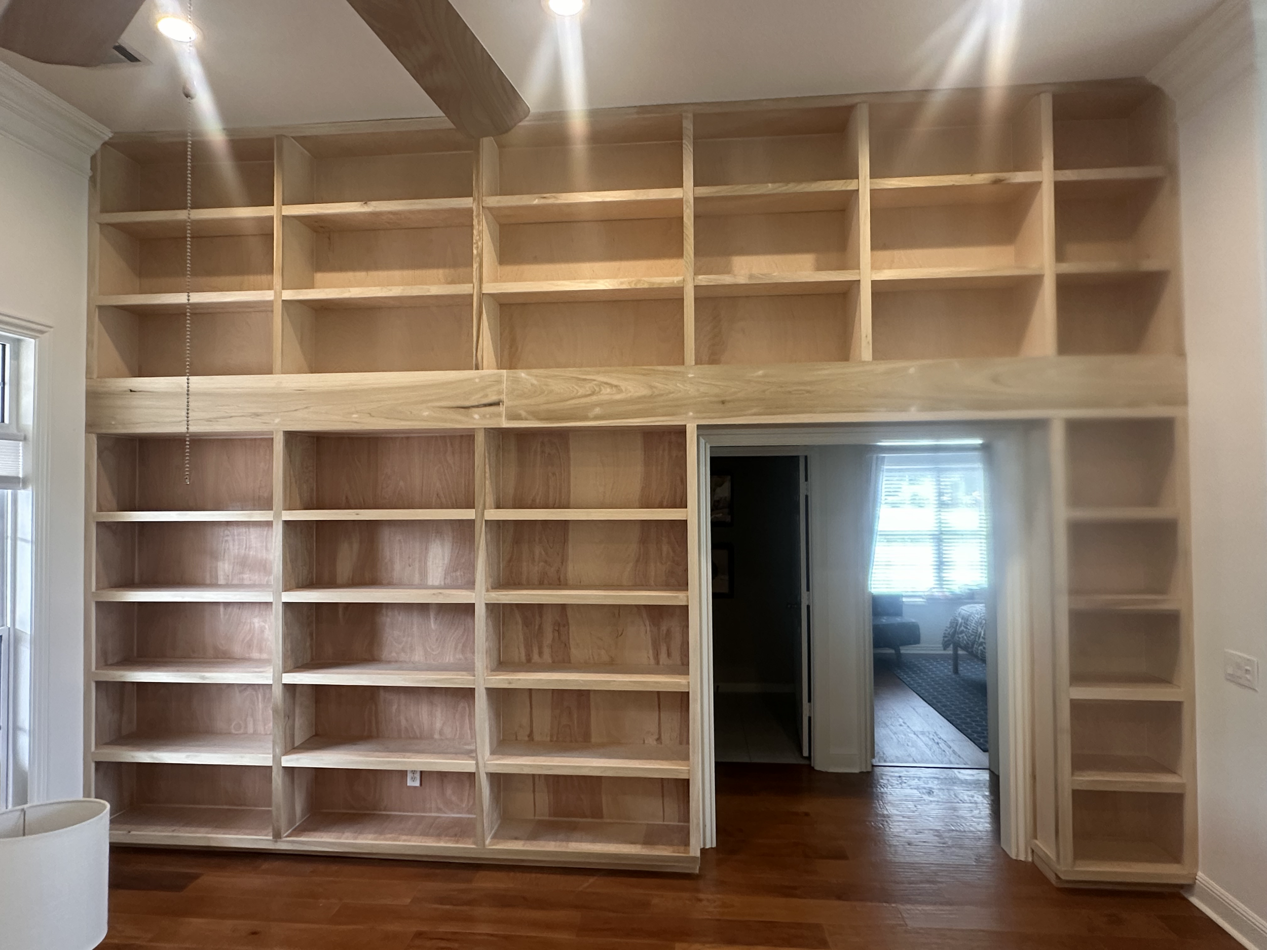 Unfinished wooden built-in bookshelves in a room, with an open doorway leading to another room with a window and bed.