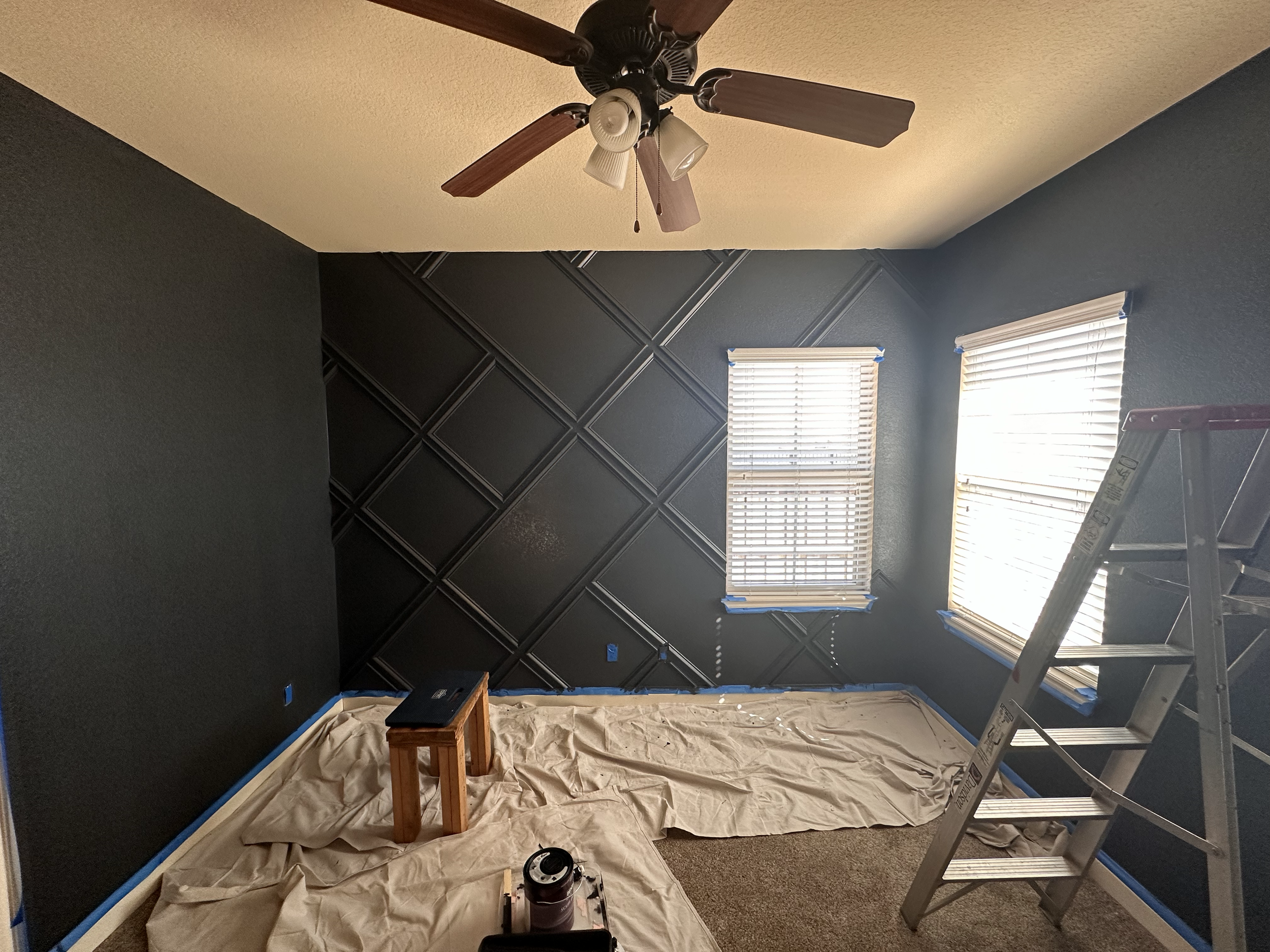 A room with dark blue walls, two windows with blinds, and a ceiling fan with lights. The wall on the left side has been painted, and the wall on the right side is partly in the process of being painted. There is a ladder on the right side, a small wo