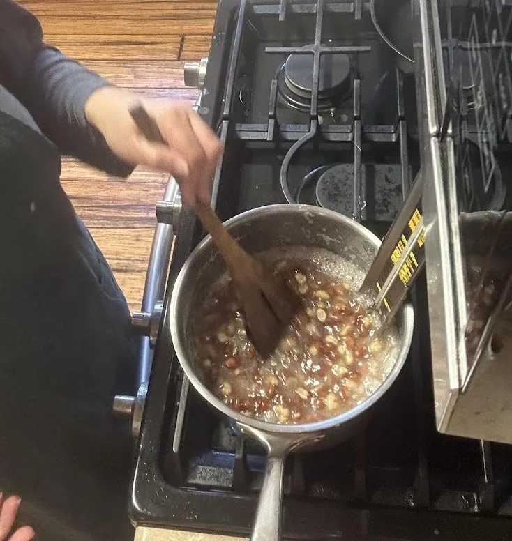 Person stirring a pot on a stovetop with a wooden spoon, containing a mixture of chopped vegetables or possibly fruit in liquid.