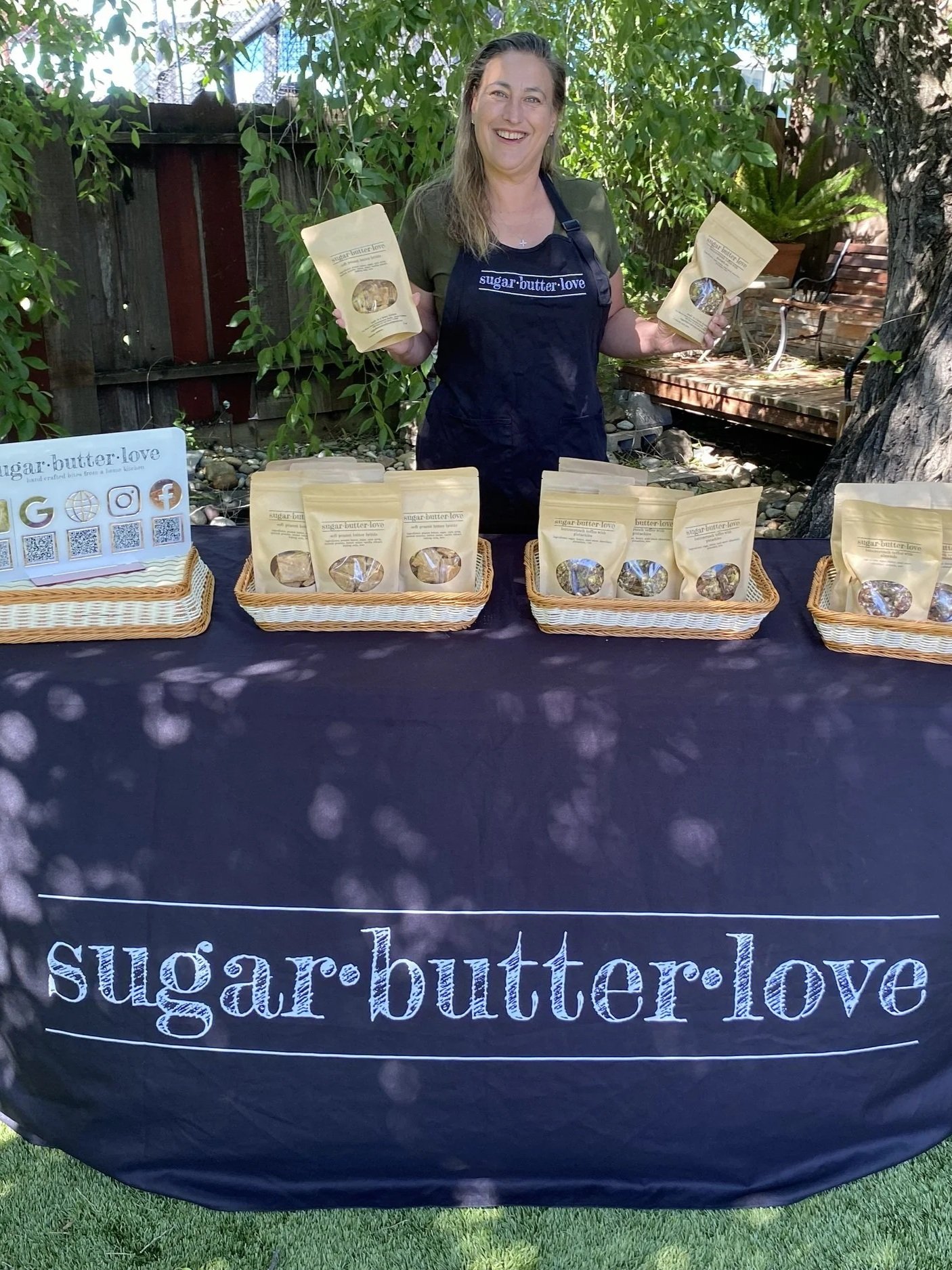 A woman stands behind a table with baskets of sugar butter love products outdoors. She is smiling, holding packages, wearing a black apron with the logo 'sugar•butter•love'. The table has a black cloth with 'sugar•butter•love' written on it, and there is a white sign with social media icons and QR codes on the left. Green foliage and a wooden fence are in the background.