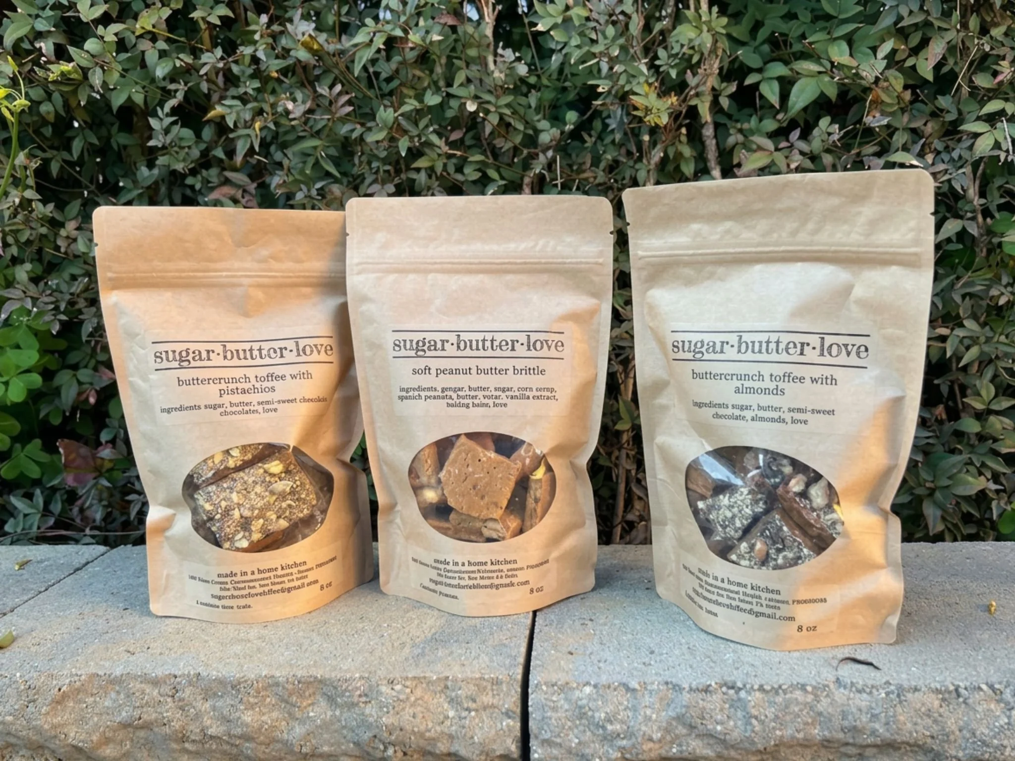 Three beige paper bags of different flavors of Sugar Butter Love toffee snacks are placed on a concrete surface in front of a bush with green leaves. The bags have transparent windows showing the toffee inside and detailed labels with flavors and ingredients.