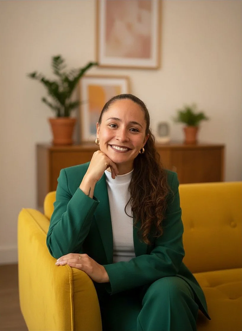 Eine lachende Frau sitzt auf einem gelben Sofa, trägt einen grünen Anzug und hat braune, lange, gewellte Haare, im Hintergrund zwei Pflanzen und Wandbilder.