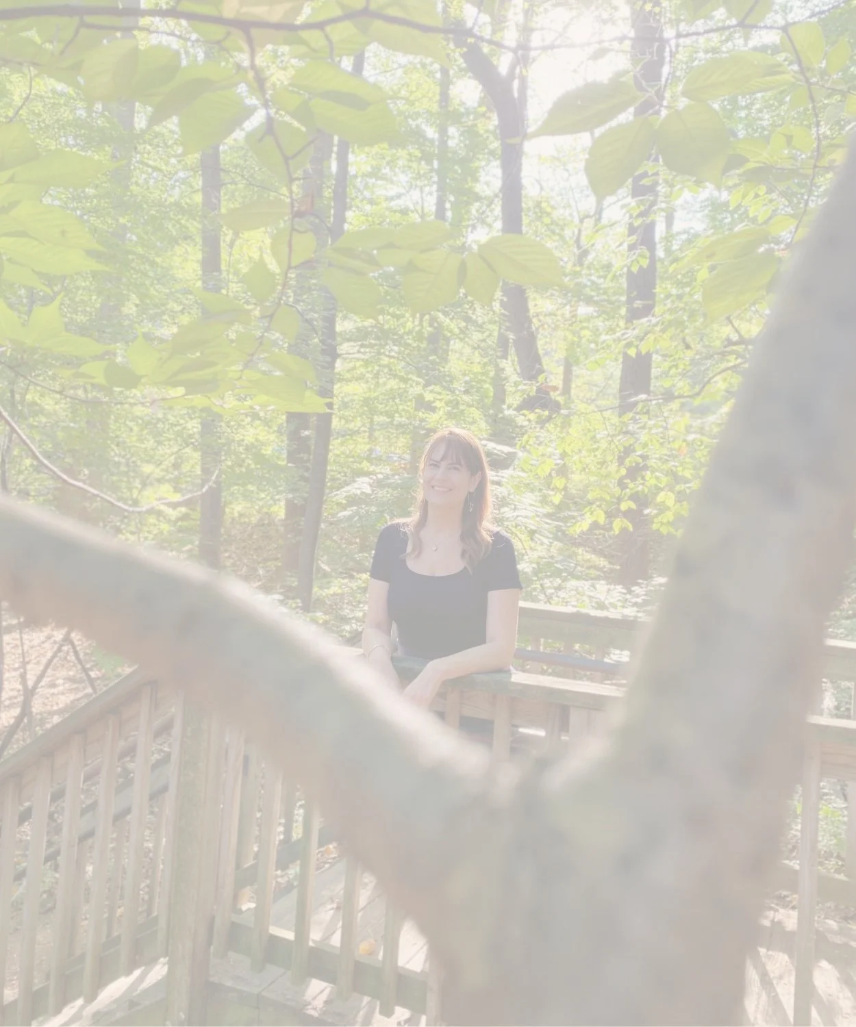 A smiling woman with brown hair and earrings, sitting on a wooden deck surrounded by trees in a forest, seen through a blurry branch in the foreground.
